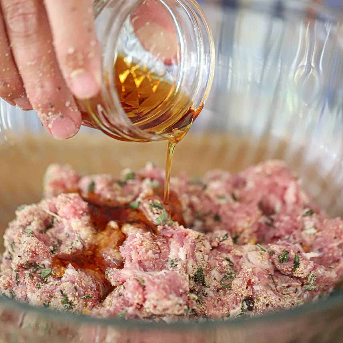 A person pouring pure maple syrup from a small glass jar into a glass bowl filled with ground pork mixed with fresh herbs.
