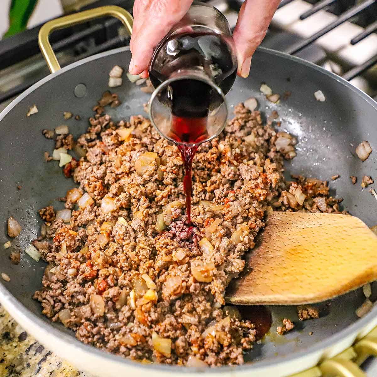 A person pouring red wine from a small carafe into a skillet filled with cooked ground beef, sautéed onions, and garlic in tomato paste.