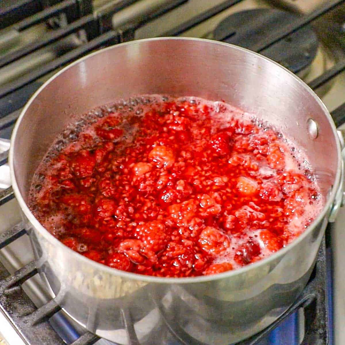 A silver saucepan filled with simmering raspberries in a boiling water with sugar on a gas stove.