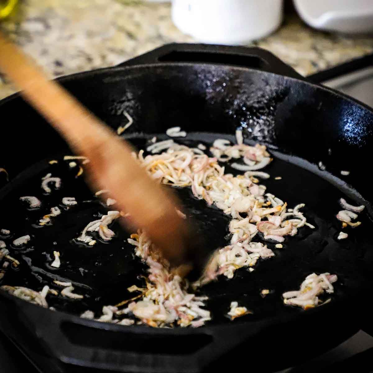 A person using a wooden spatula to stir chopped shallots that are being sautéed in a large cast-iron skillet on a gas stove.