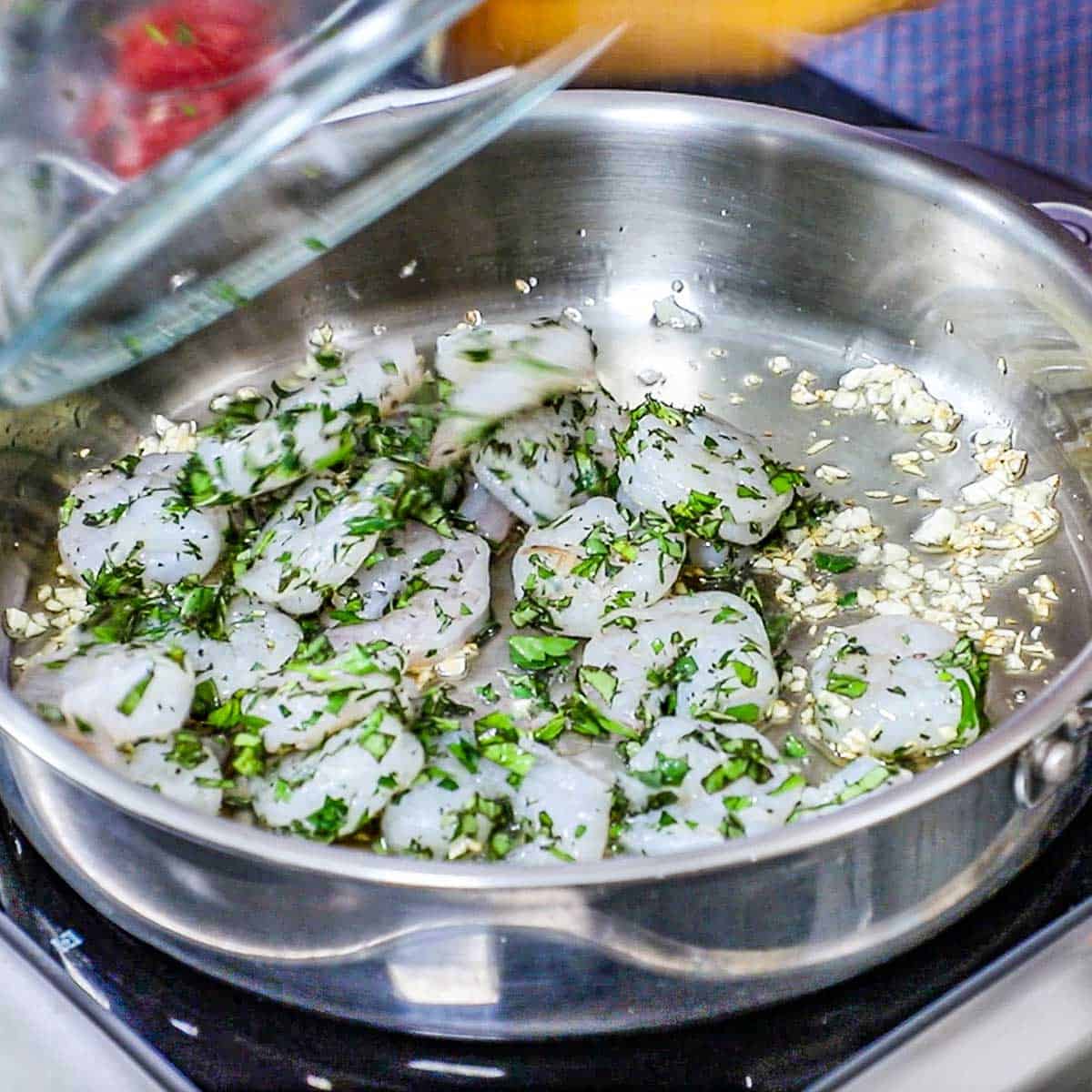 A person transferring seasoned uncooked shrimp into a large skillet that is filled with sautéed mince garlic in oil.