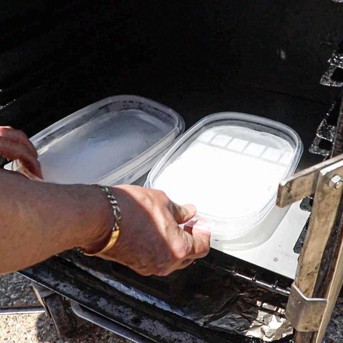 A person placing two plastic containers filled with a block of ice onto a cold smoke plate that is resting on the bottom grate of an electric smoker.