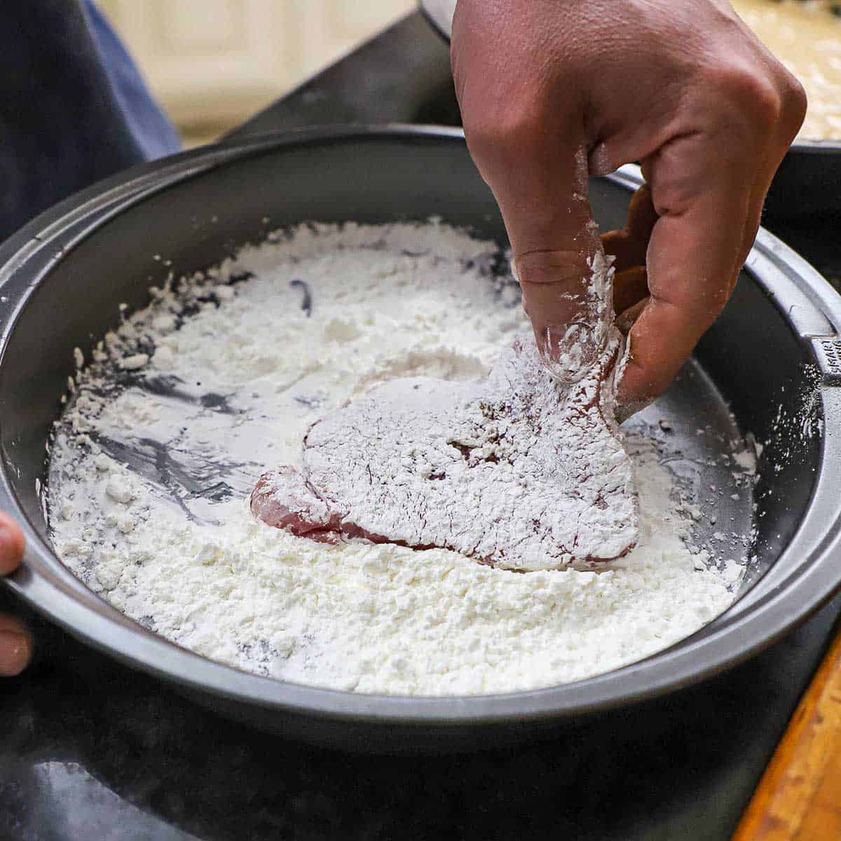 A person using his hand to dredge a seasoned boneless, thin pork chop through flour in a round baking pan.