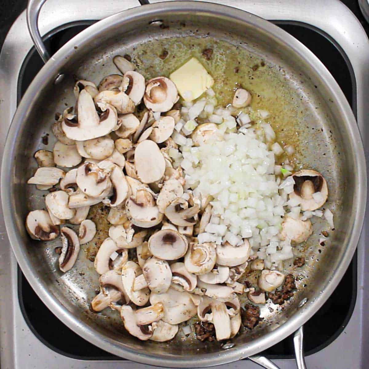An overhead view of a large stainless steel skillet filled with sliced mushrooms and chopped onions being sautéd in butter.