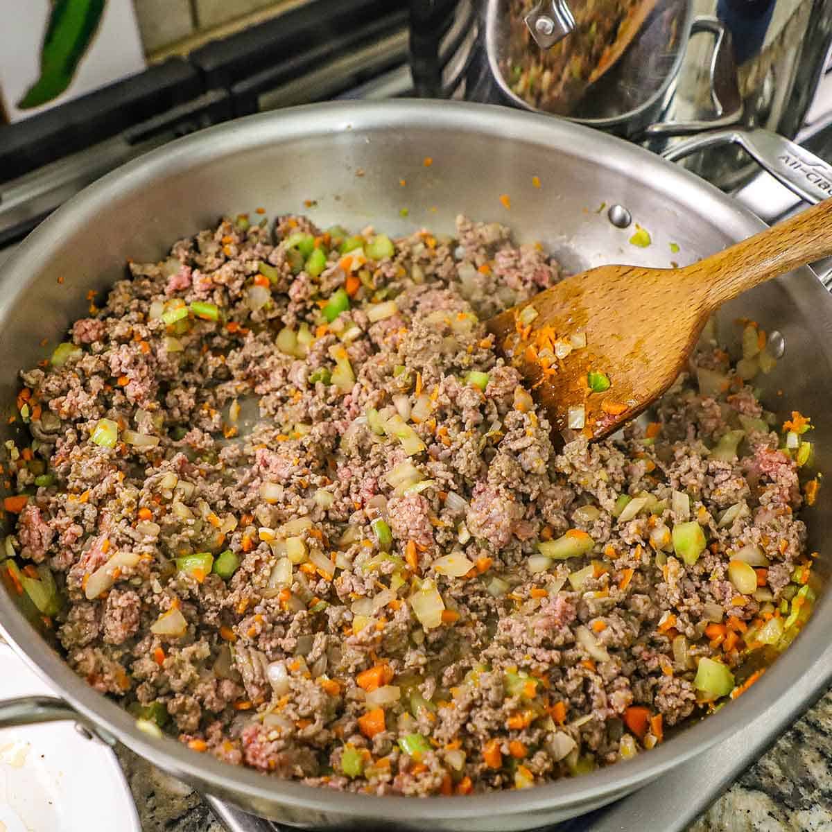 A large silver skillet filled with cooked ground beef and Italian sausage simmering with chopped onion, carrots, and celery.