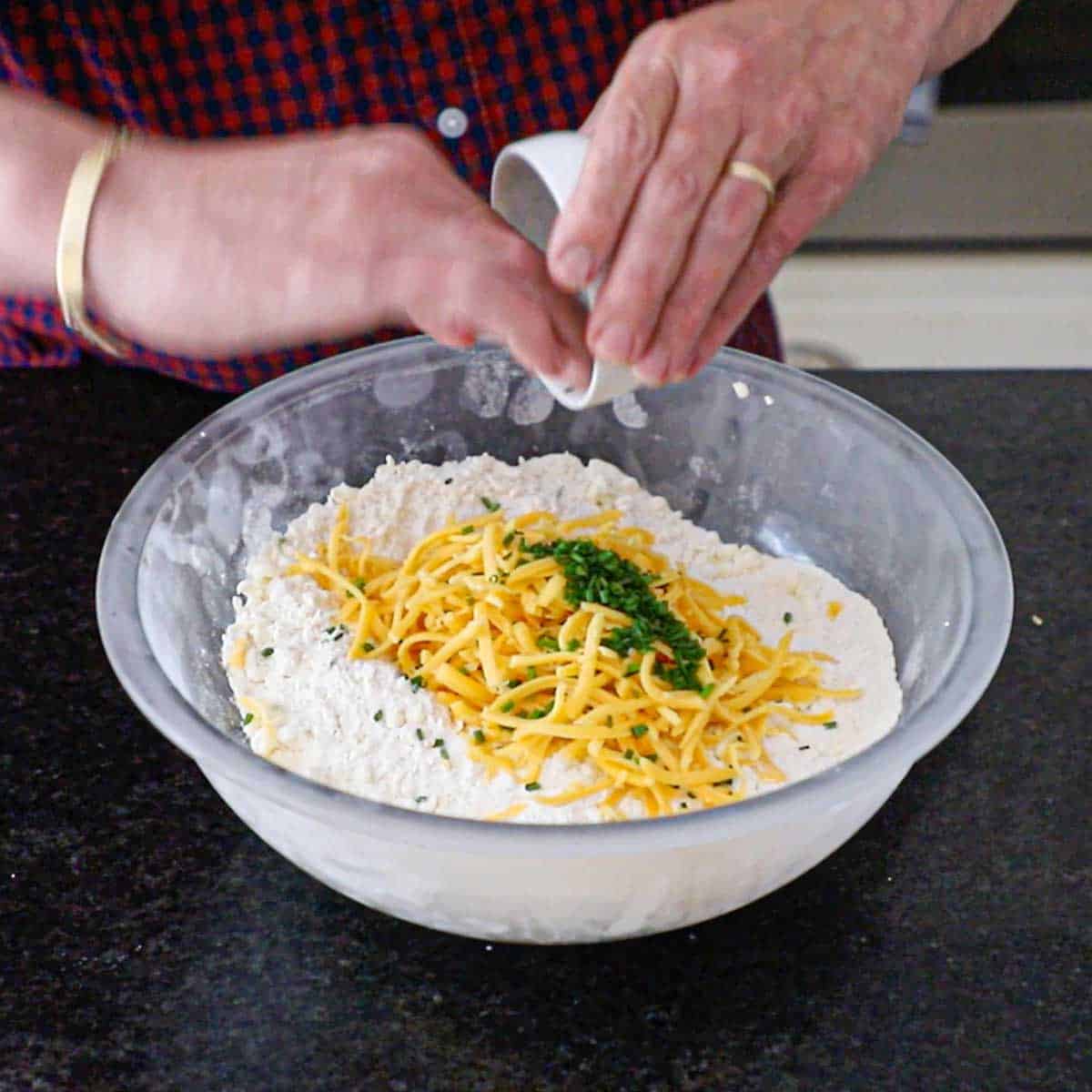 A person using his fingers to transfer snipped chives from a small bowl into a large glass bowl containing flour mixed with chilled grated butter and topped with shredded cheddar cheese.