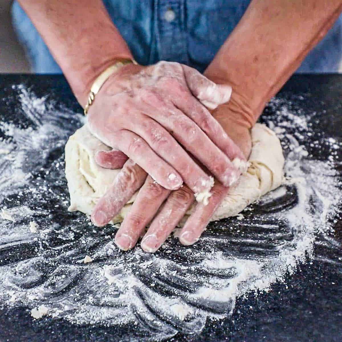 A person using the palms of his hands to knead homemade naan dough on a floured marble countertop.