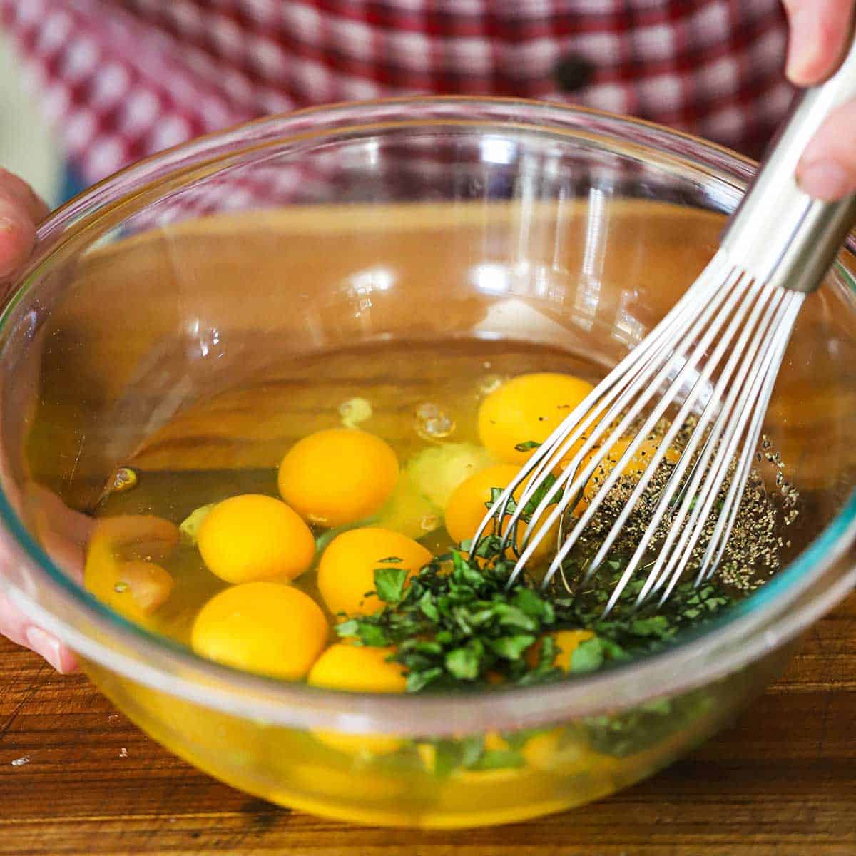 A person using a whisk to combine 12 large eggs with chopped fresh basil, salt, and pepper in a large glass bowl.