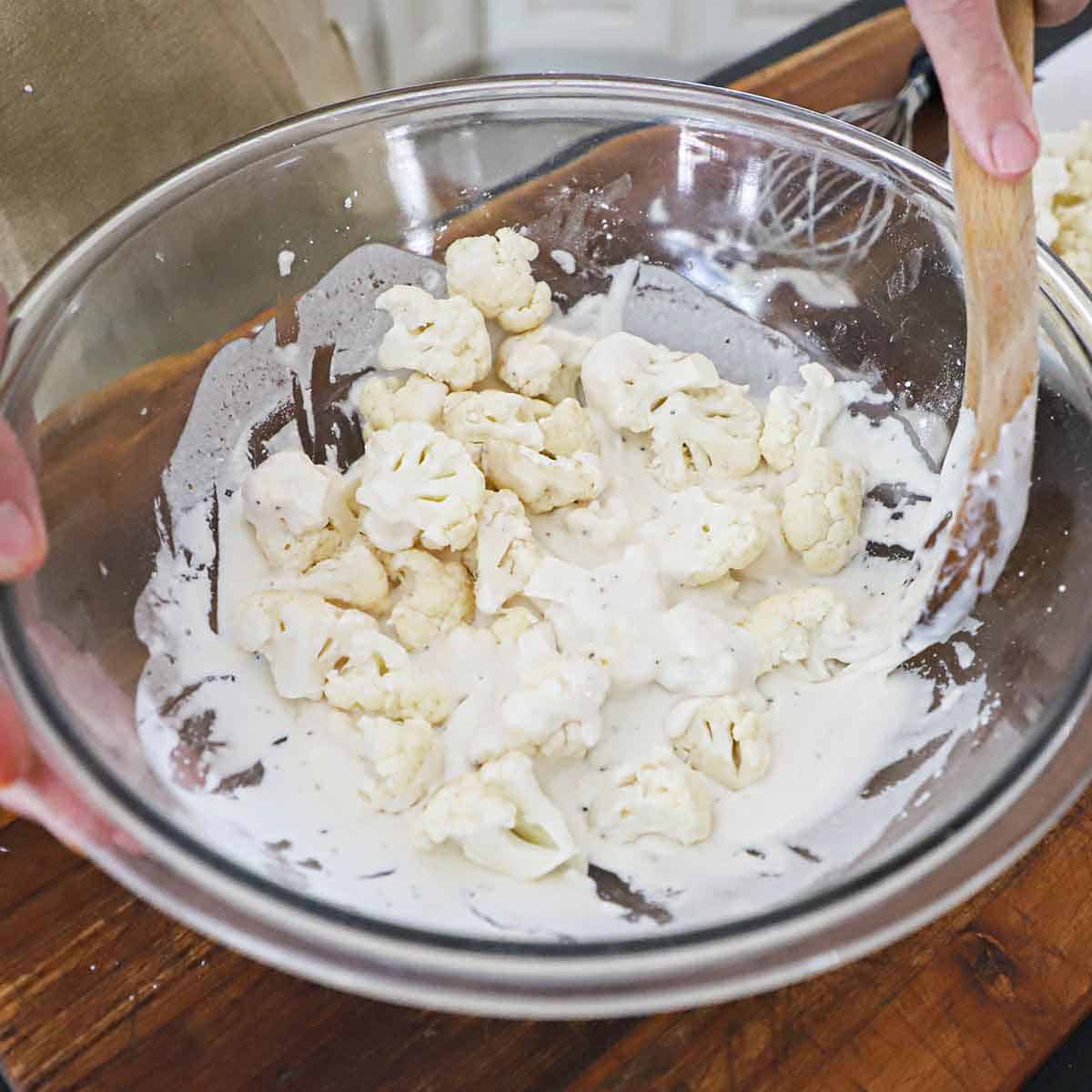 A person using a large wooden spoon to stir cauliflower florets in a large glass bowl filled with a white batter for deep-frying.