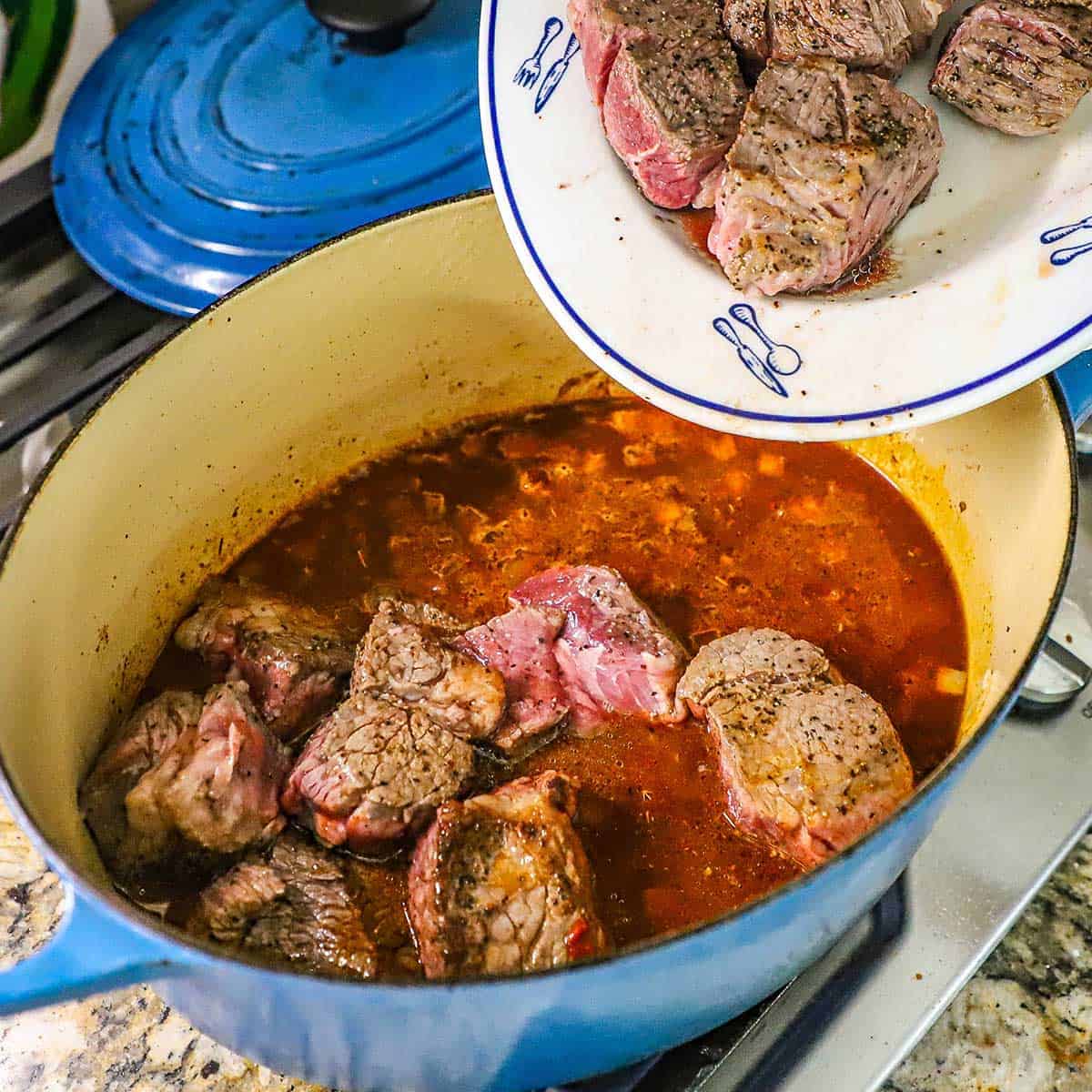 A person transferring seared chunks of chuck roast into a simmering broth of beef broth, tomatoes, onions, and seasonings.