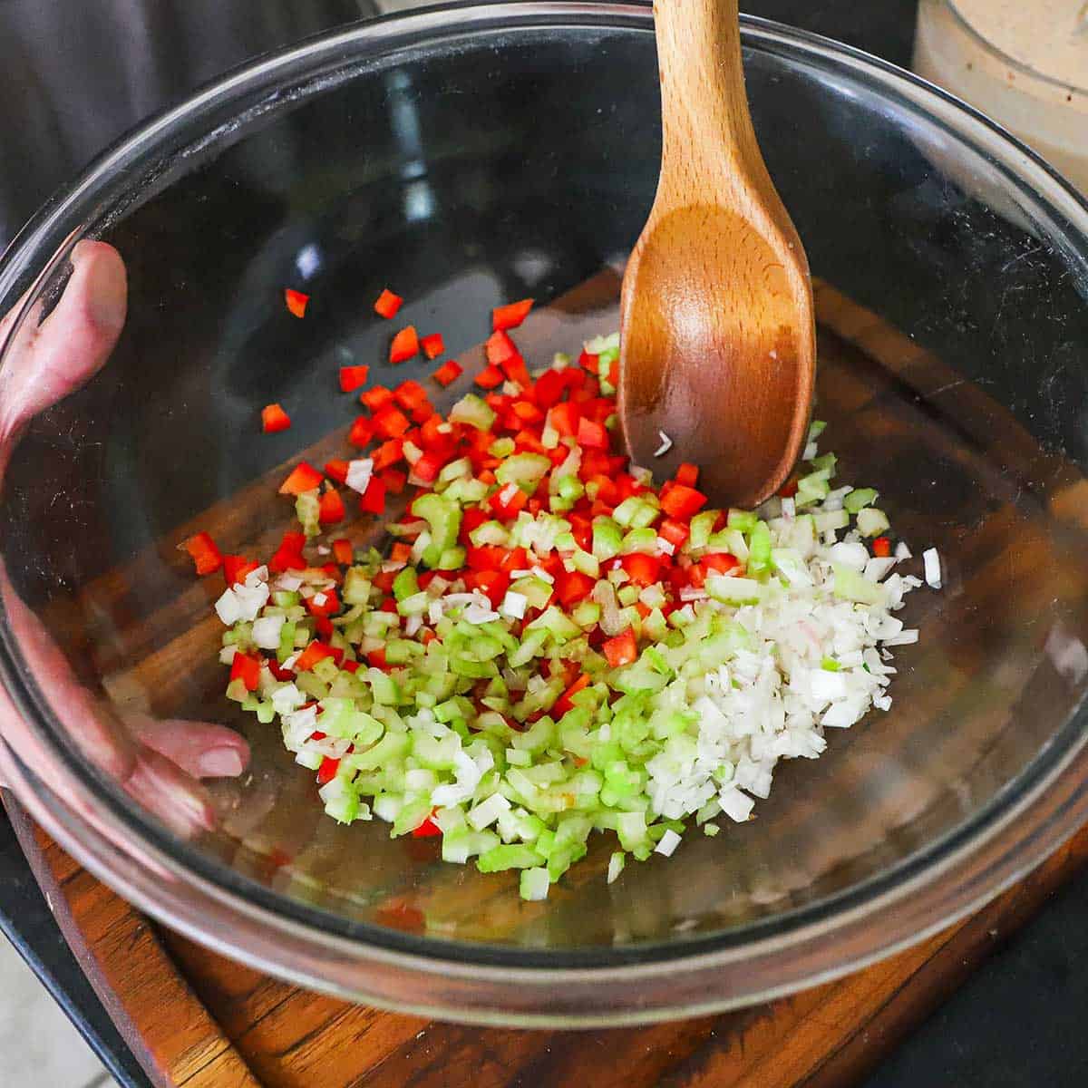 A person using a wooden spoon to stir chopped celery, red bell pepper, and shallots together in a large glass bowl.