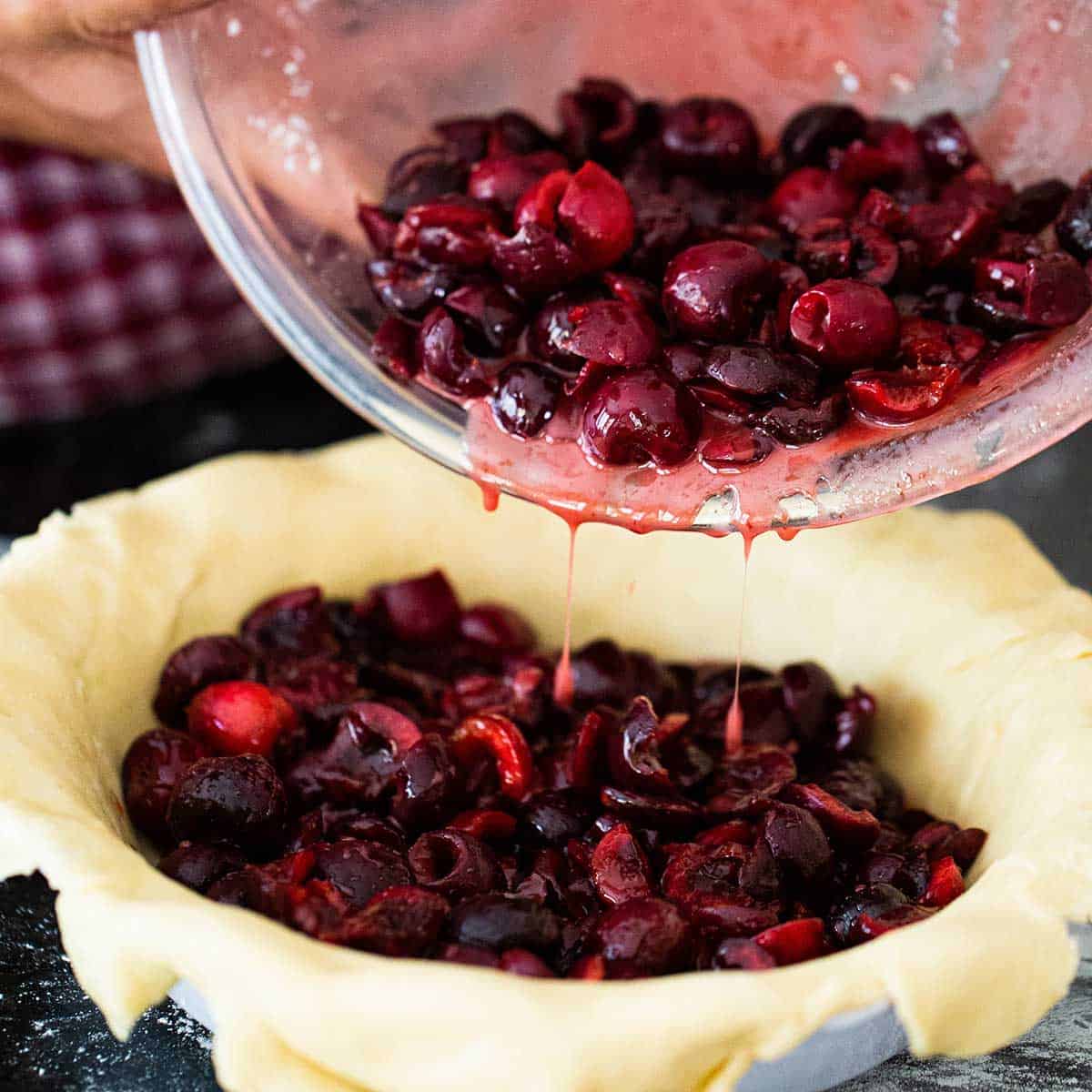 A person transferring a homemade cherry filling from a large glass bowl into a pie dish that is lined with homemade pie dough.