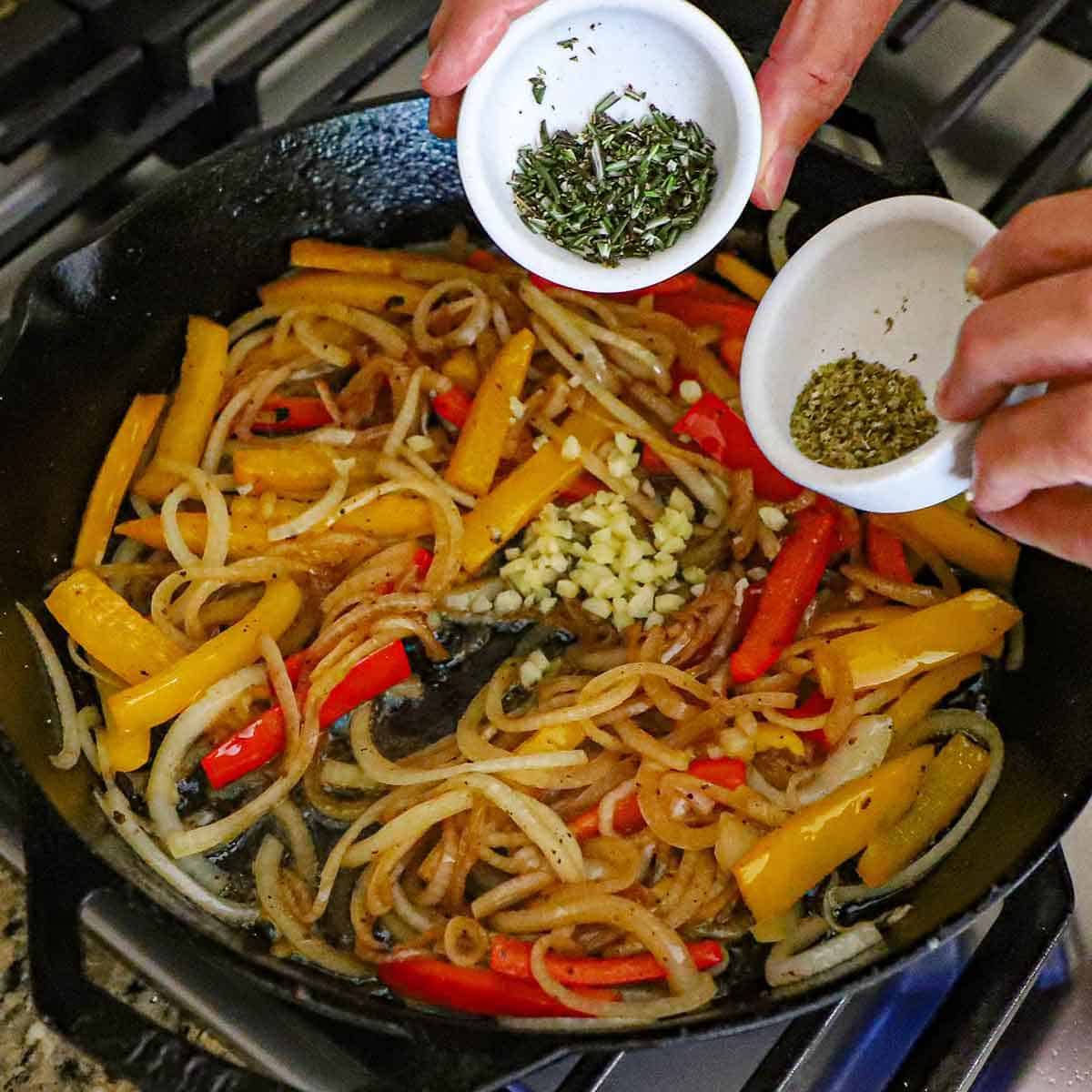 A person holding a small white bowl filled with chopped fresh rosemary in one hand and the other hand holding another small bowl filled with dried oregano both over a skilet filled with sautéed onions and strips of bell peppers.