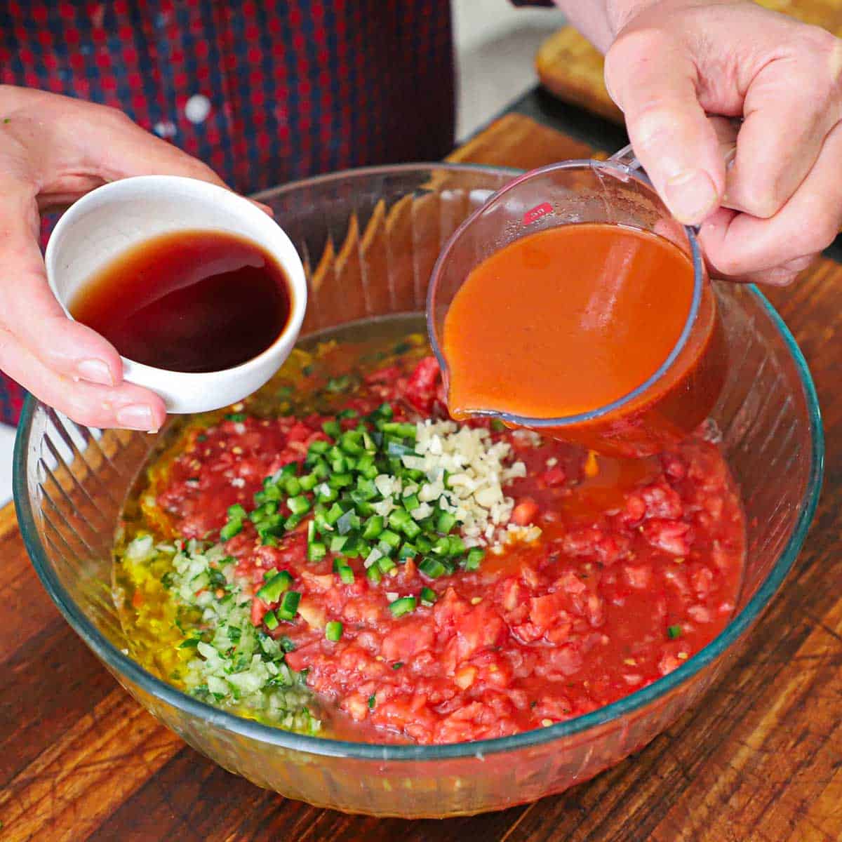 A person holding a small white bowl filled with red wine vinegar in one hand and a measuring cup filled with tomato juice both over a large glass bowl filled with all the components of classic gazpacho.