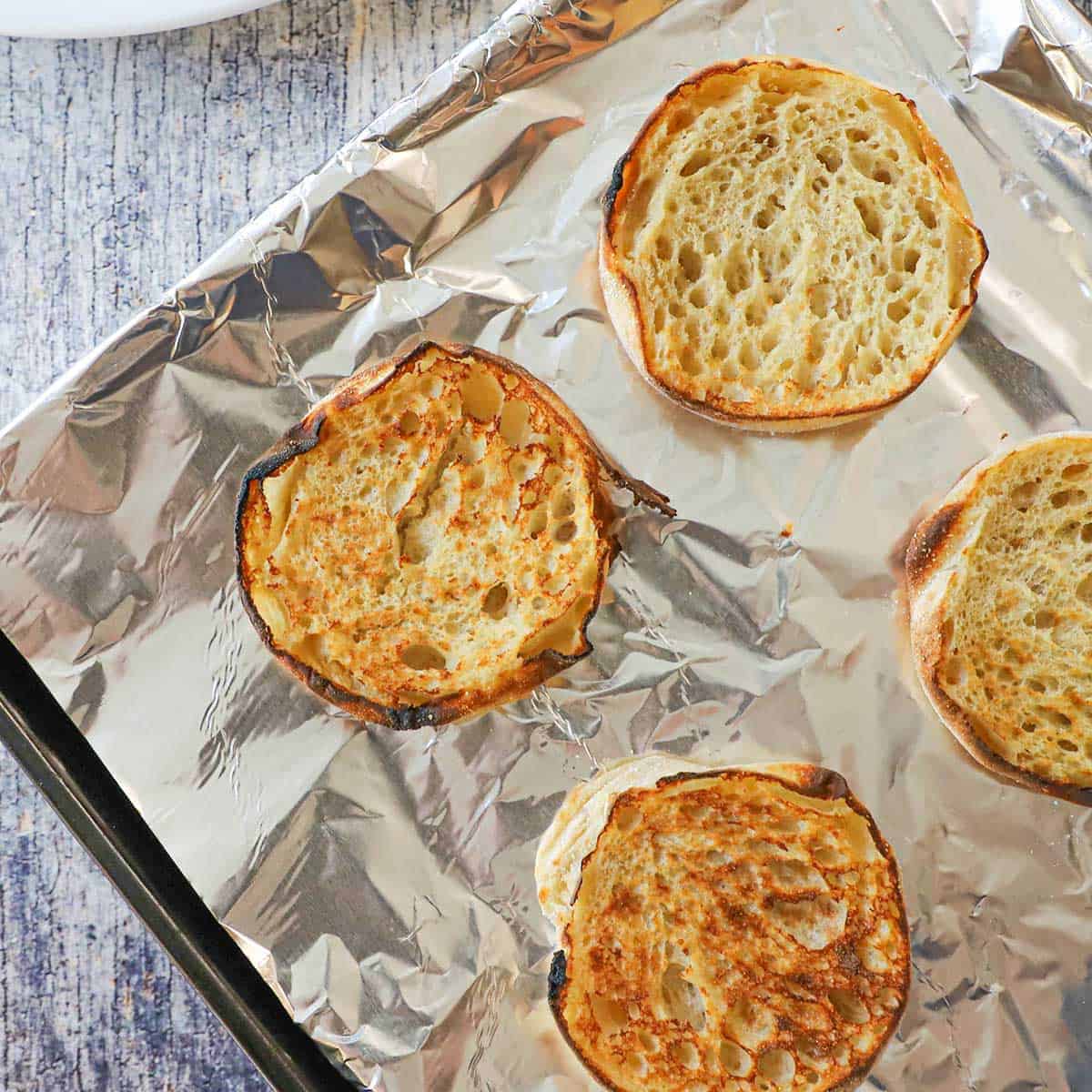 An overhead view of four English muffins that have been split in half and have been toasted on a baking sheet lined with aluminum foil.