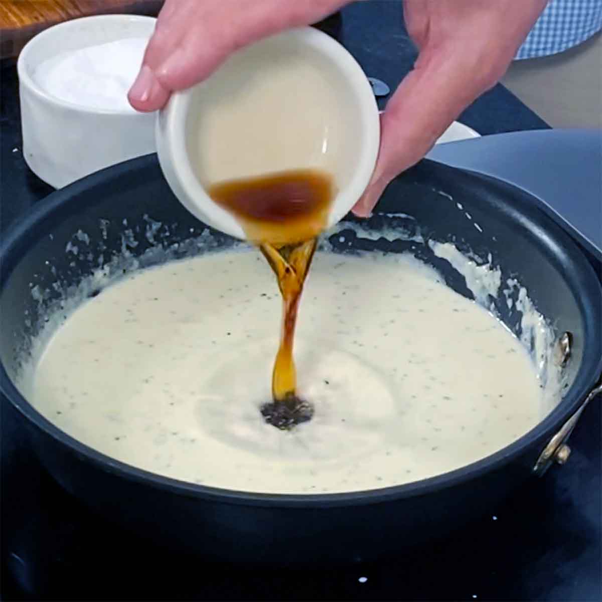 A person pouring a small amount of Sherry vinegar into a small skillet filled with a simmering garlic-infused cream sauce.