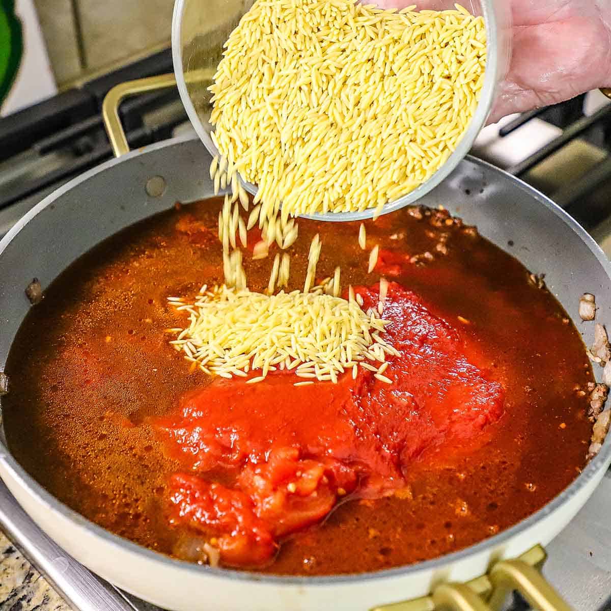 A person transferring a small bowl of uncooked orzo pasta into a large skillet filled with simmering beef stock, crushed tomatoes, and a meat sauce.