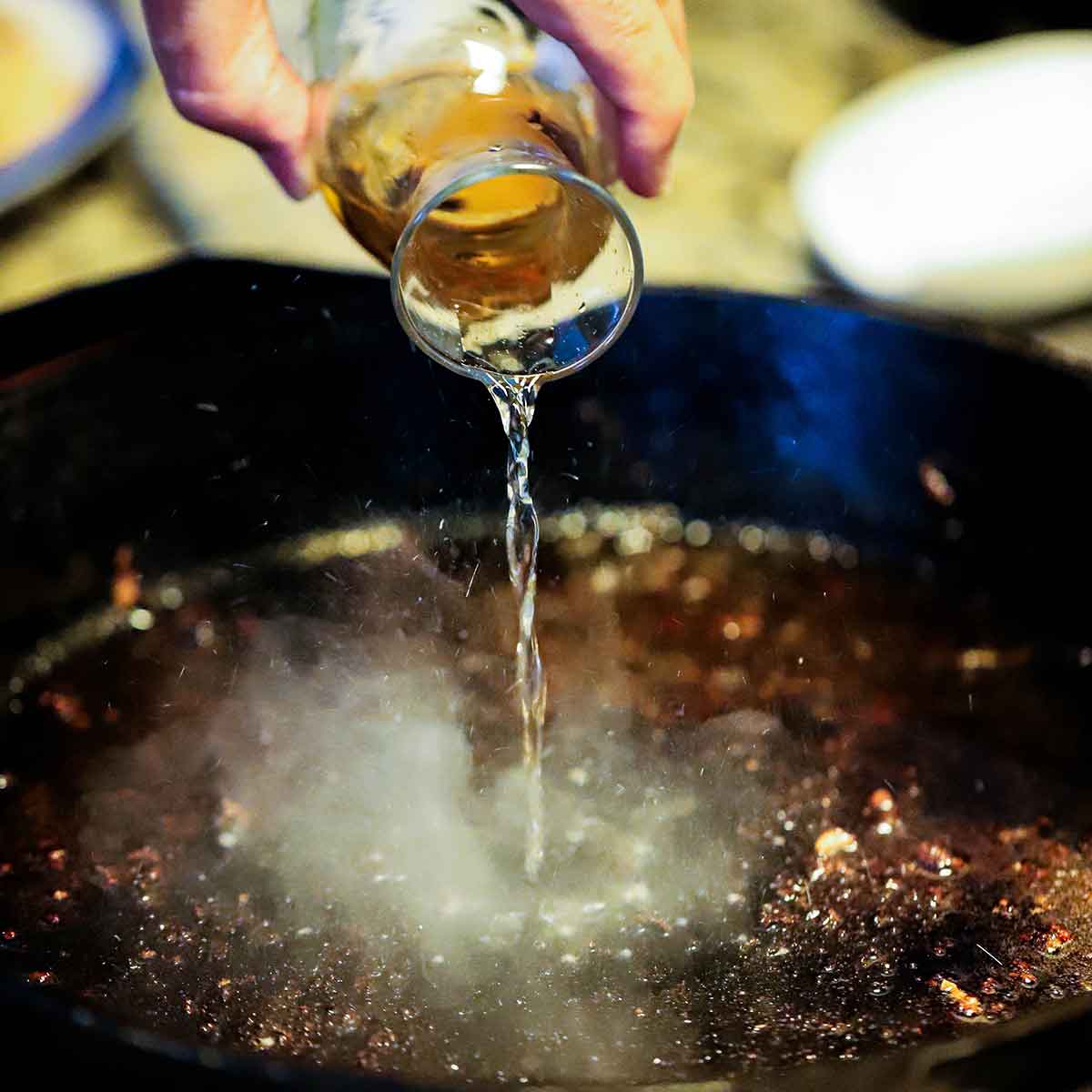 A person pouring Sherry wine from a small glass carafe into a large cast-iron skillet over heat causing the wine to sizzle.