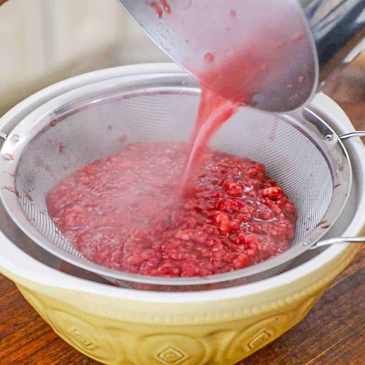 A person pouring simmered fresh raspberries and boiling sugar water through a fine-mesh sieve into a large ceramic bowl on a wooden cutting board.