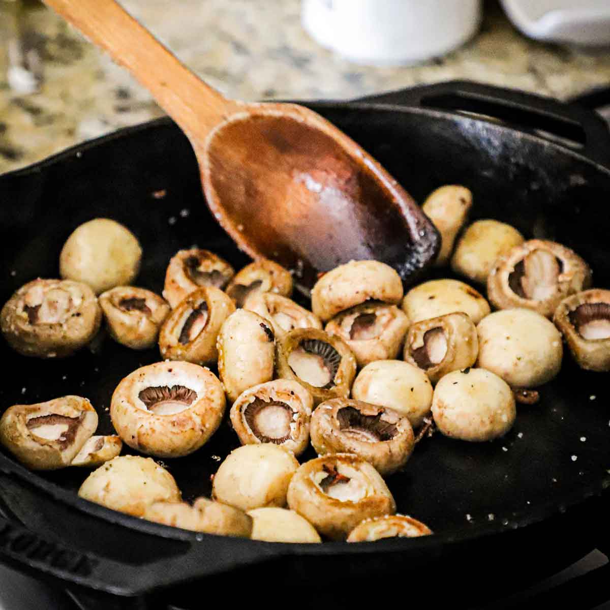 A person using a large wooden spoon to stir and sauté whole button mushrooms in olive oil in a large cast-iron skillet on a gas stove.