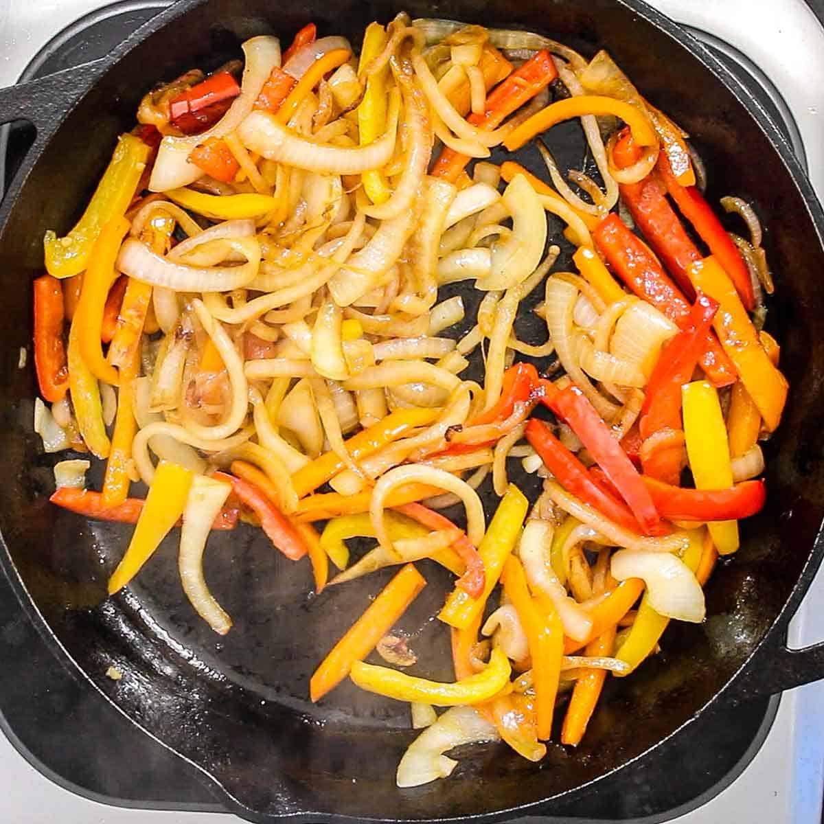 An overhead view of strips of onion, red bell pepper, and yellow bell pepper being sautéed in oil in a large cast-iron skillet.