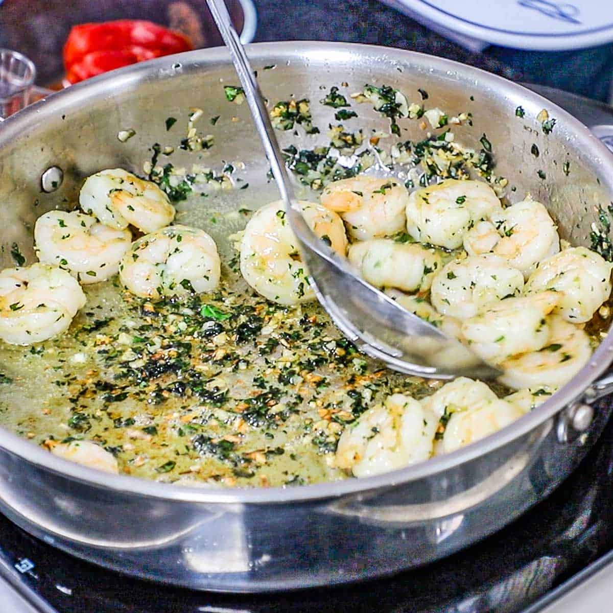A person using a large slotted spoon to removed cooked shrimp from a large skillet filled with more of the shrimp, as well as sautéed garlic and fresh herbs.