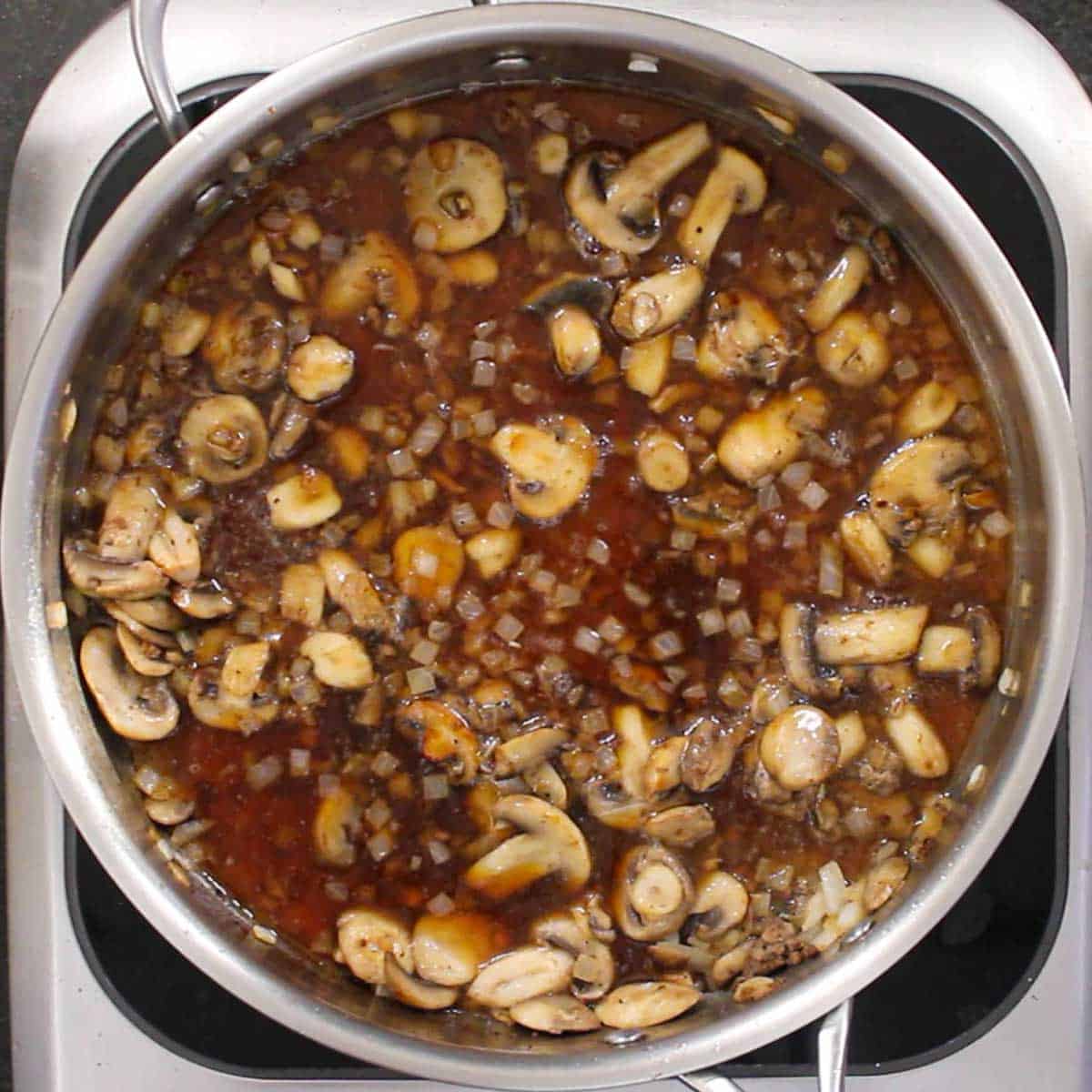 An overhead view of sautéed sliced mushrooms and chopped onions simmering in beef broth in a large skillet.