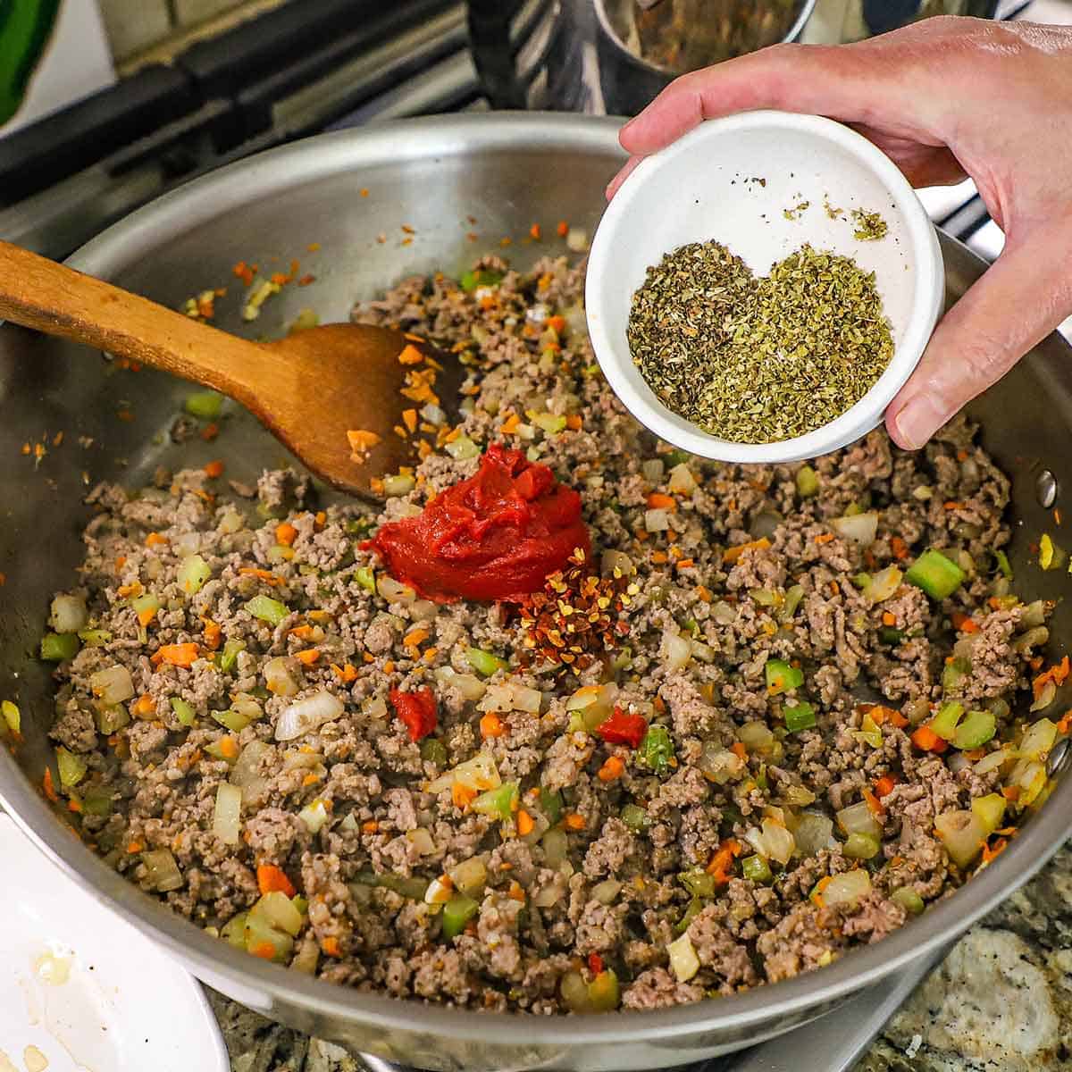 A person holding a small bowl filled with dried oregano and basil over a skillet filled with simmering ground beef and sausage, mirepoix, and a blob of tomato paste.