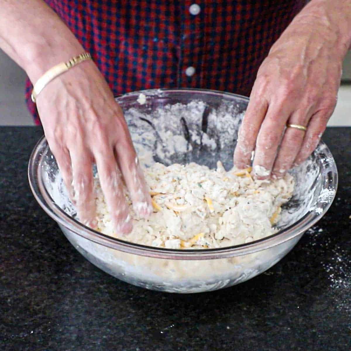 A person using both of his hand to mix cheddar biscuit dough in a glass bowl into a shaggy dough.