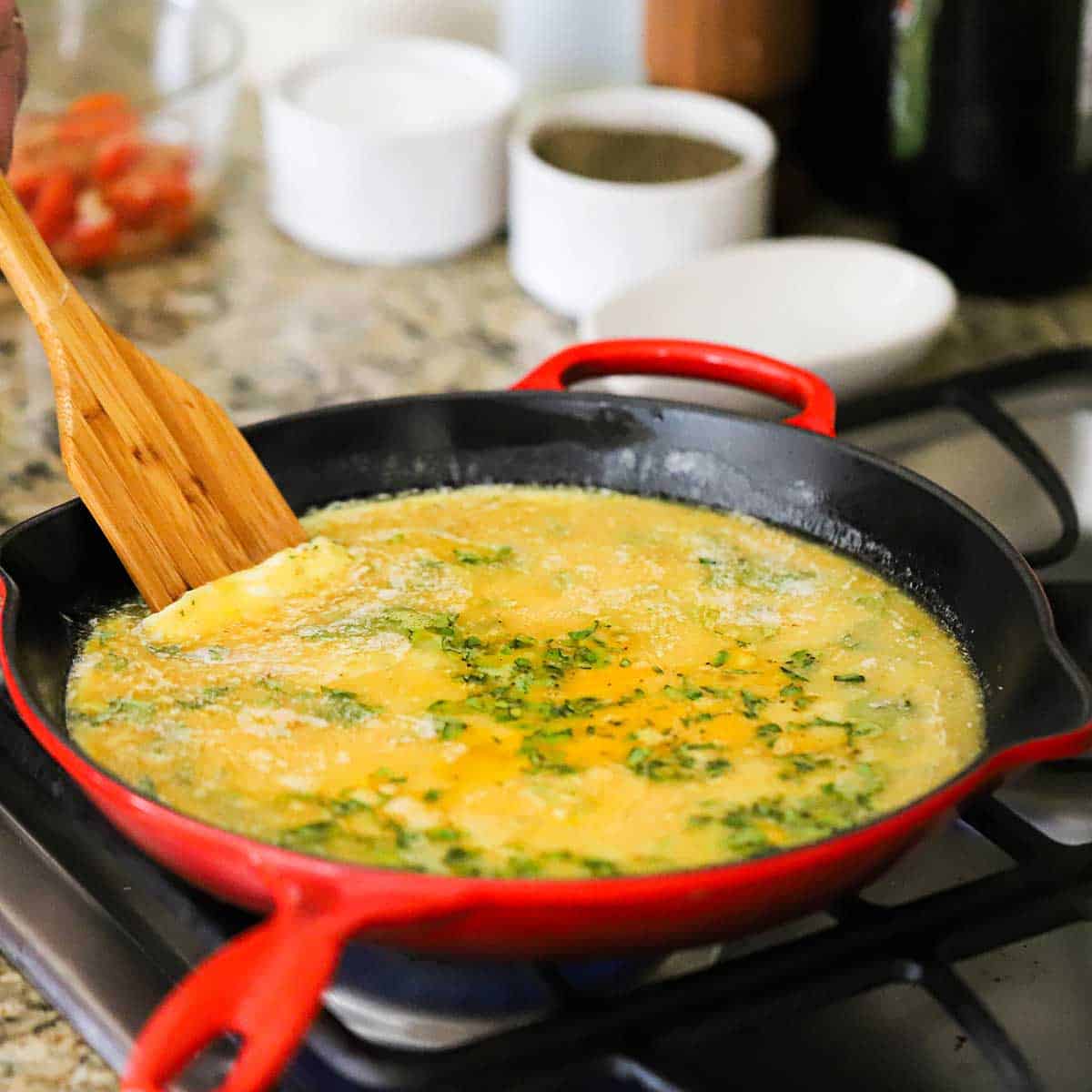 A person using a wooden spatula to scrape up scramble eggs that have begun to set in a large ceramic skillet on a gas stove.