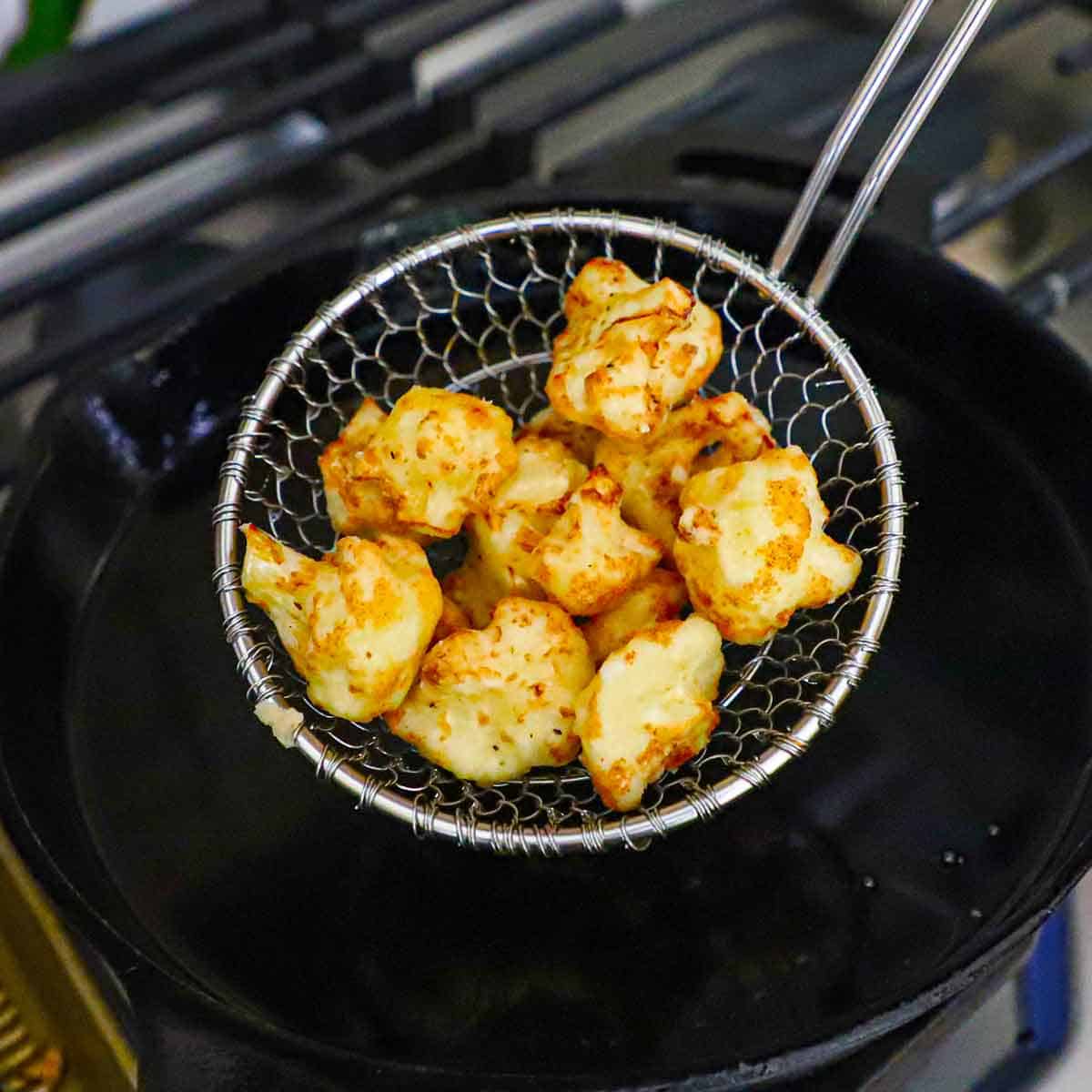 A person holding up a small pile of battered and deep-fried cauliflower florets over a skillet filled with hot oil.