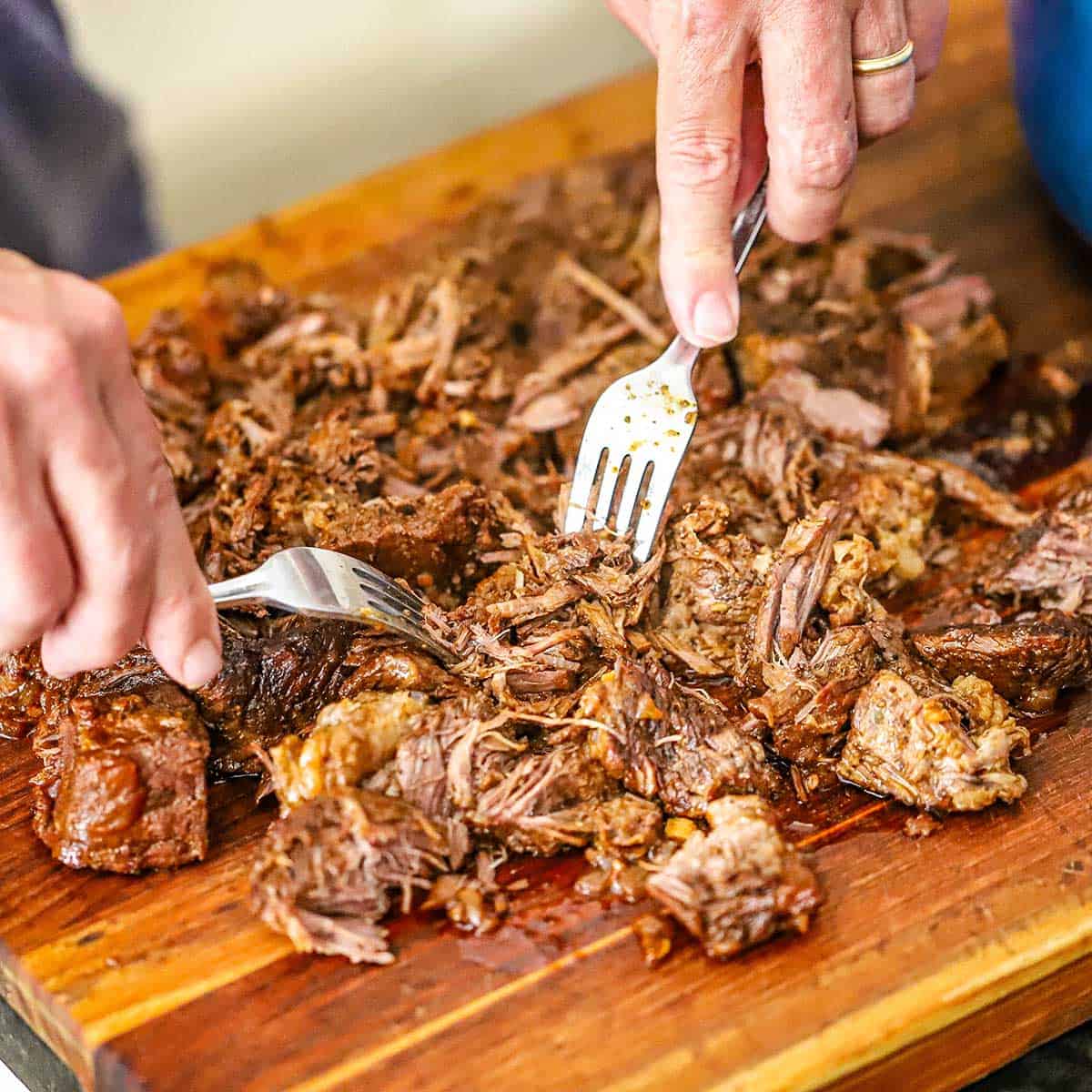 A person using a pair of forks to shred fall-apart-tender braised beef on a wooden cutting board.