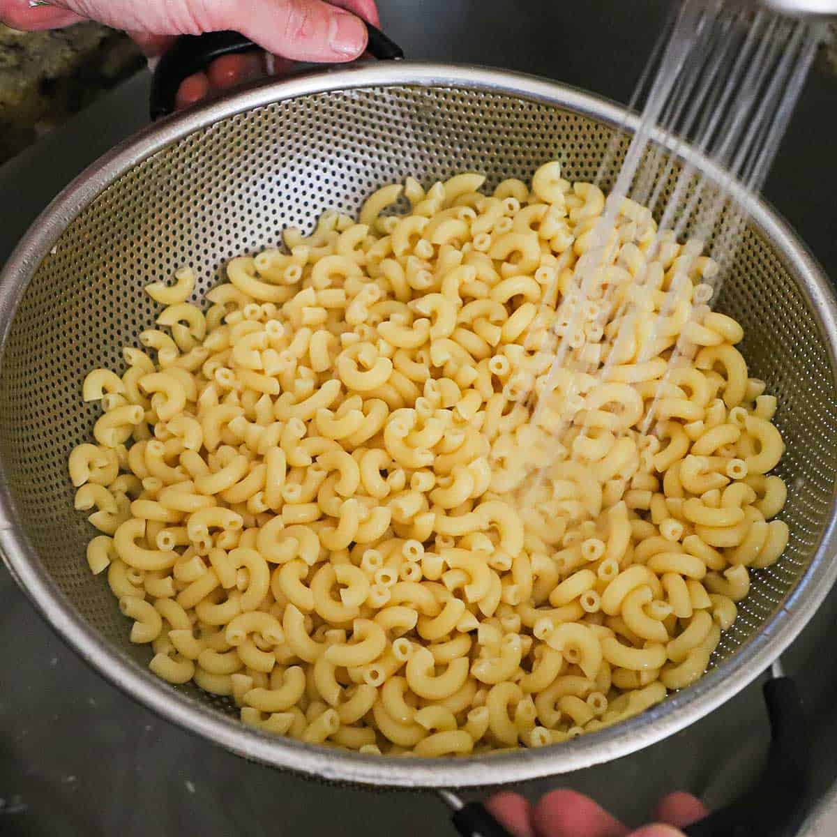 A person rinsing cooked macaroni pasta in a colander with water in a sink.