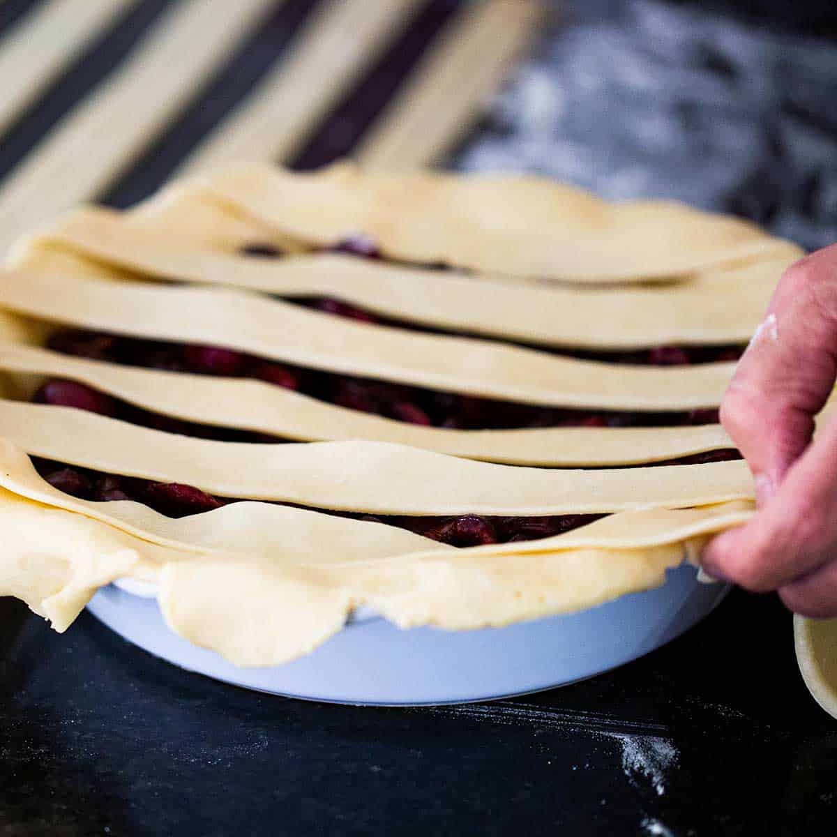 A person placing a strip of pie dough over the top of an uncooked cherry pie along with other strips of dough that are parallel with each other.