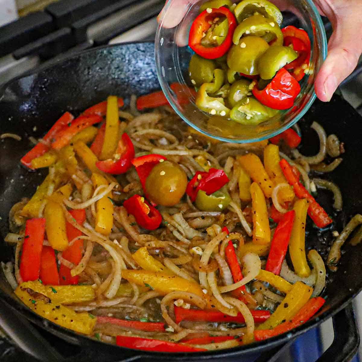A person transferring roughly chopped sweet pickled cherry peppers from a small glass bowl into a large cast-iron skillet filled with simmering sautéed onions and strips of bell peppers.