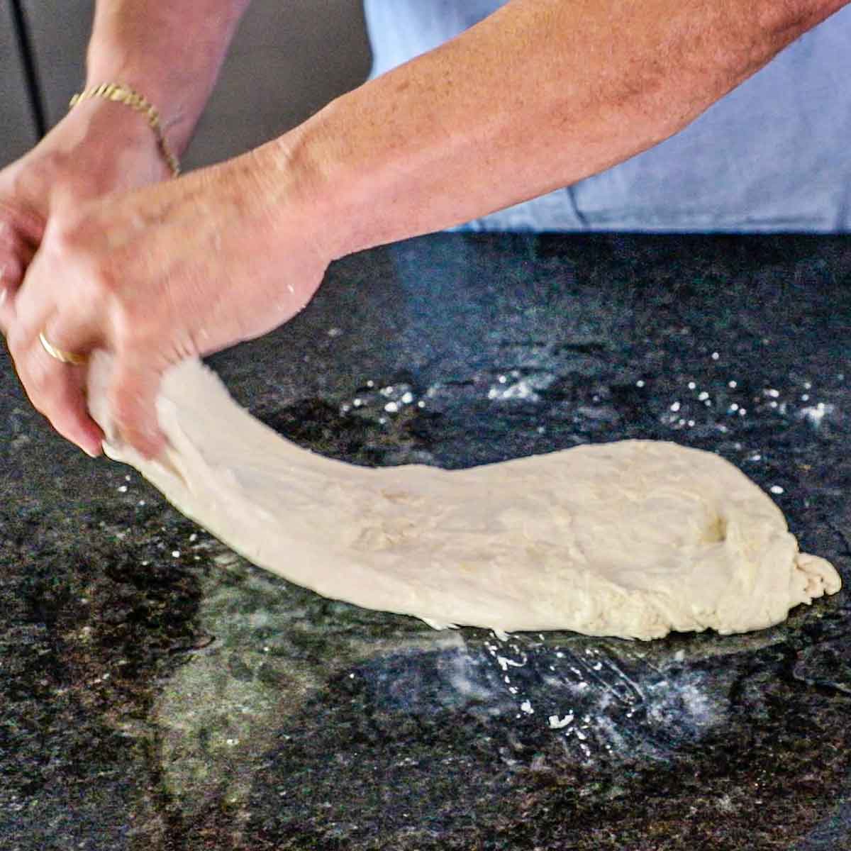 A person using his hands to stretch one end of a rectangular piece of highly hydrated dough on a black marble counter that is wet with water.