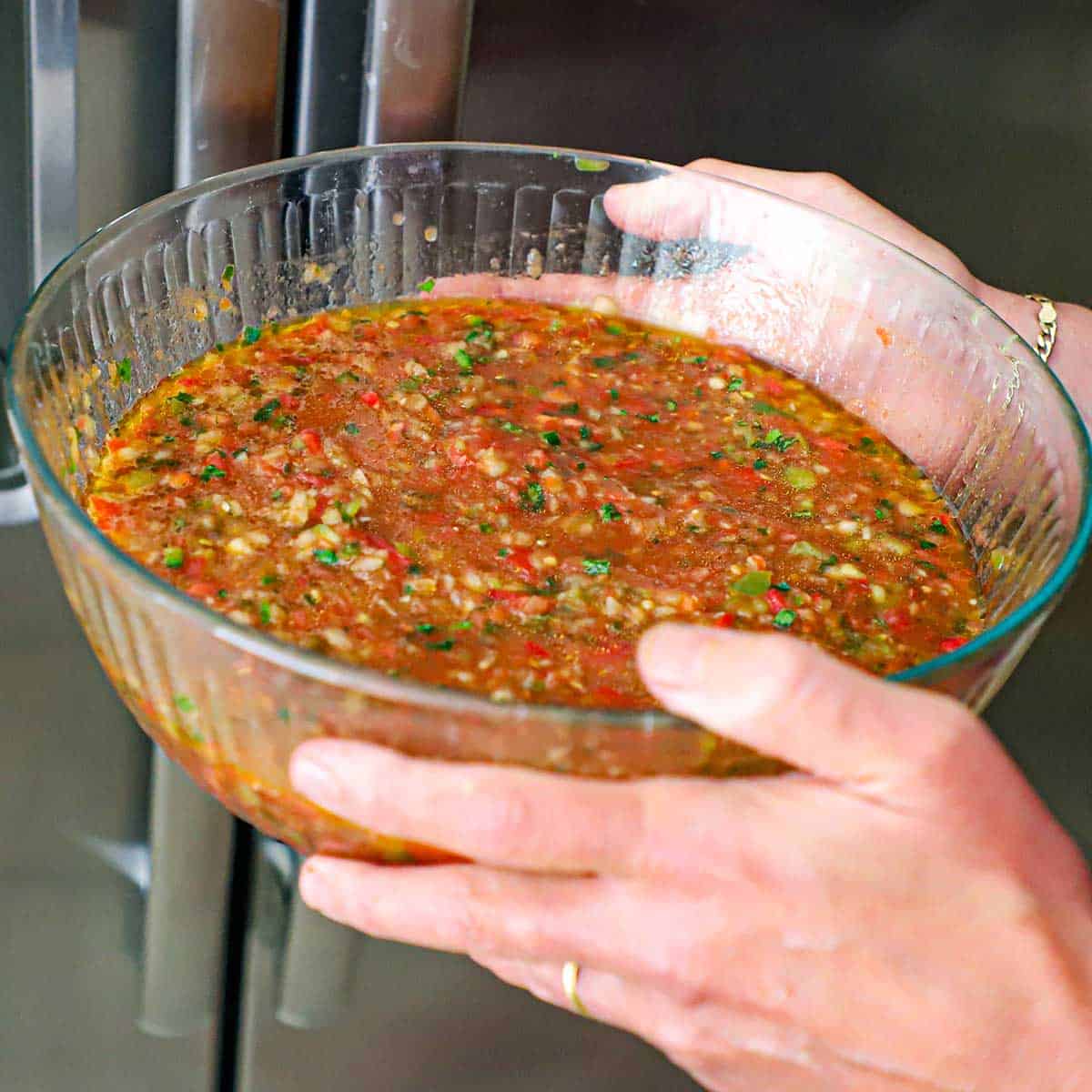A person holding large glass bowl filled with classic gazpacho that he is getting ready to place inside a refrigerator.