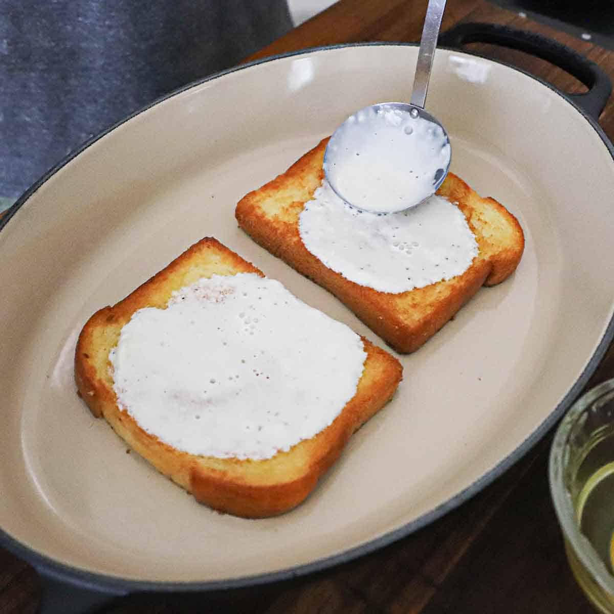 A person using a small ladle to slather on a garlic-infused cream sauce over two slices of toasted Brioche bread sitting side by side in an oval baking dish.