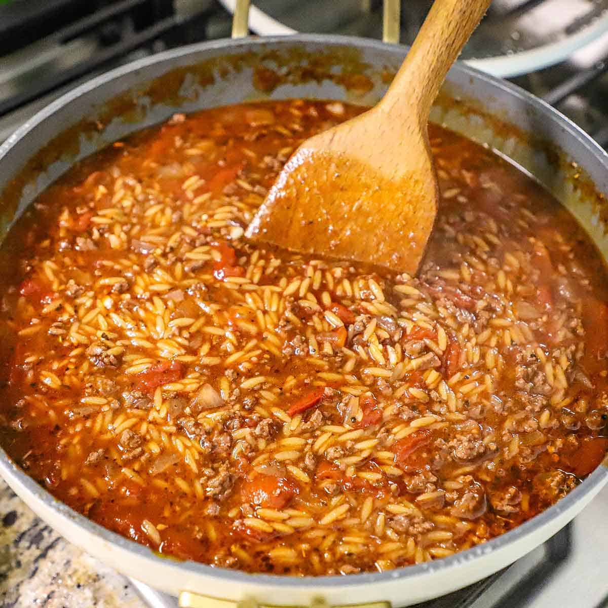 A person using a large wooden spatula to stir partially cooked orzo pasta in a large skillet that is filled with a ground beef and tomato sauce.