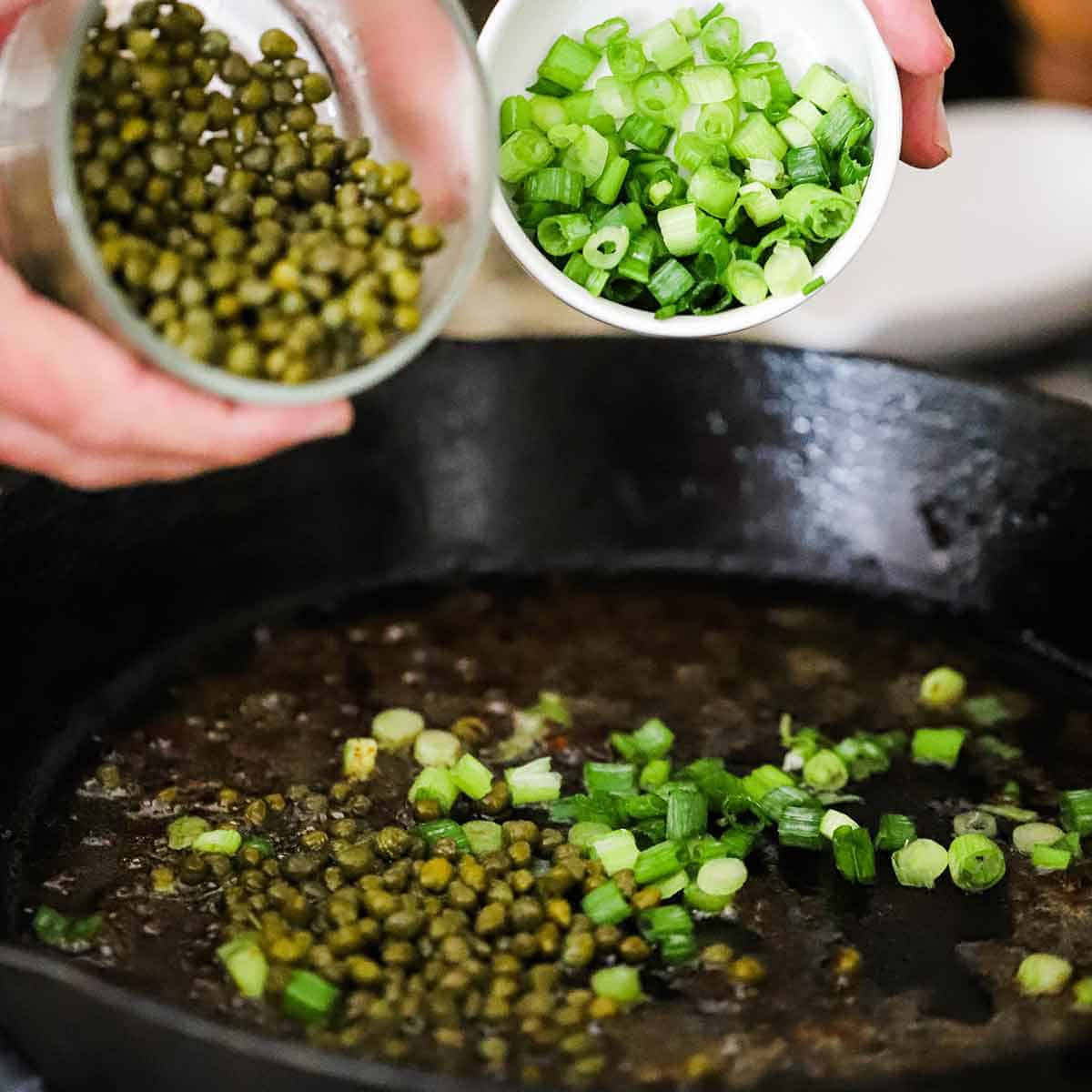 A person transferring capers from a small bowl into a cast-iron skillet filled with reduced Sherry as well as transferring chopped scallions from another bowl into the same skillet.