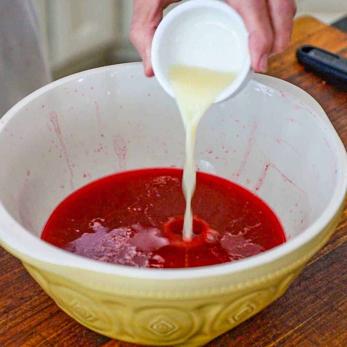 A person pouring lemon juice from a small white bowl into a large ceramic bowl partially filled with a fresh strained raspberry liquid.