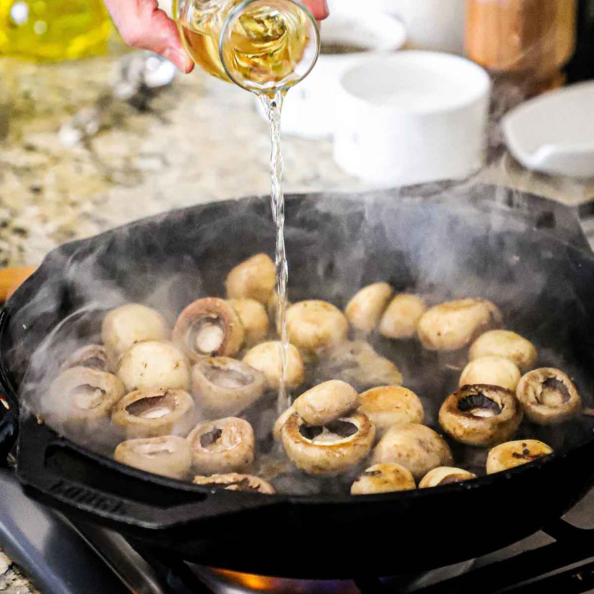 A person pouring Sherry wine from a small carafe into a large black cast-iron skillet that is filled with whole sautéed button mushrooms.