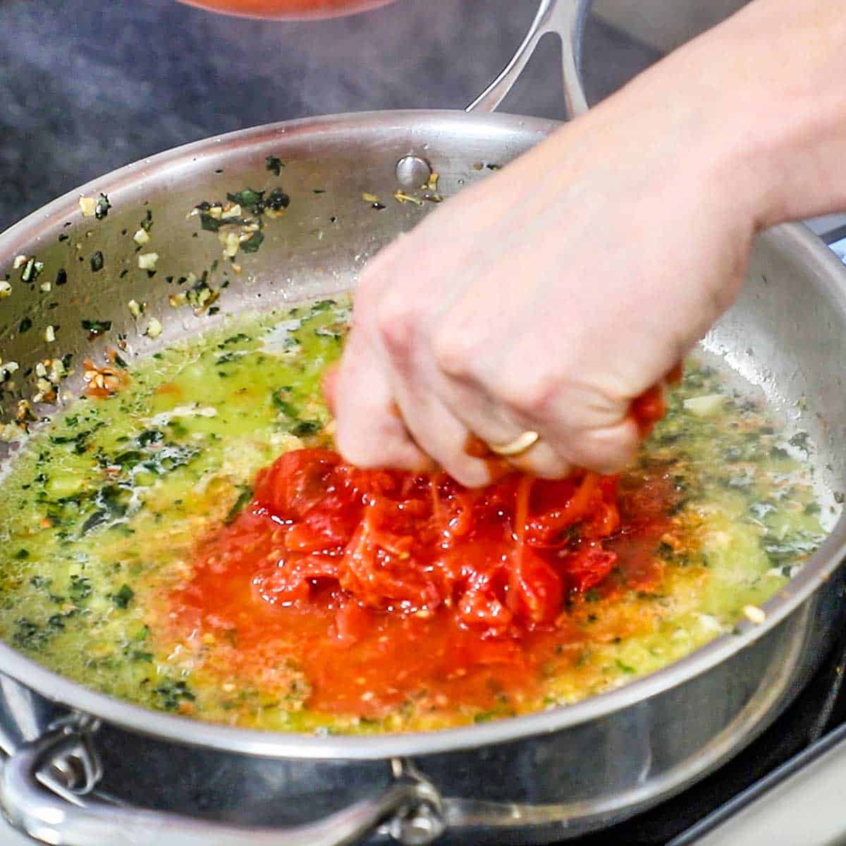 A person using his hand to crush whole San Marzano tomatoes into a large skillet filled with a simmering white wine sauce with garlic and fresh herbs.