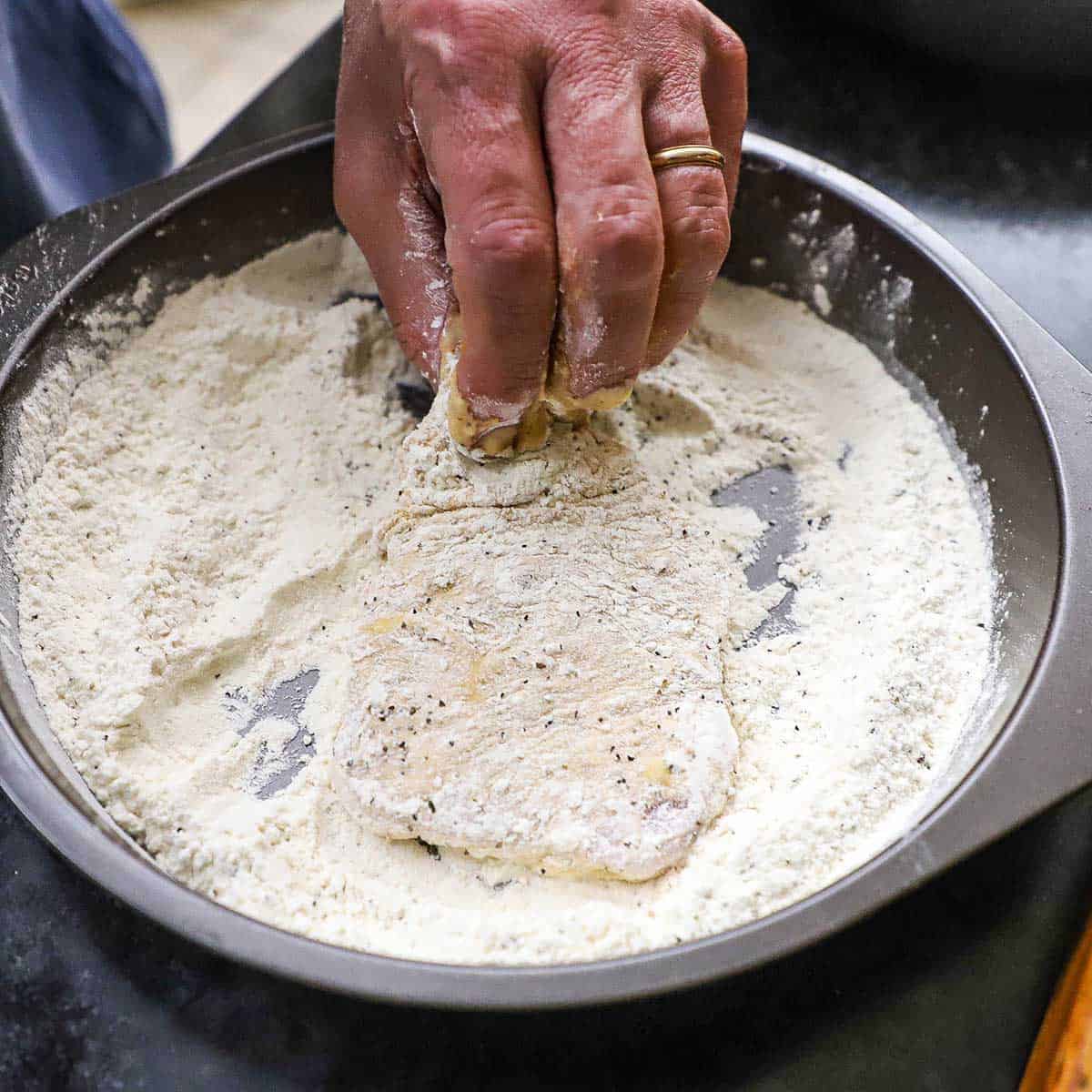 A person dredging a thin, boneless pork, that has been floured and dipped in a wet batter through a second coating flour.
