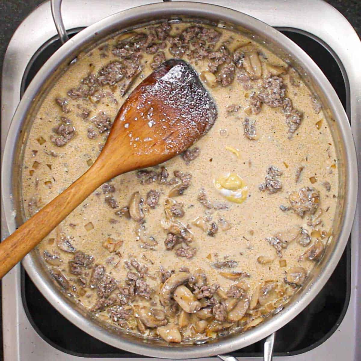 A person using a large wooden spatula to stir a creamy and beefy broth along with ground beef, sautéed mushrooms, and chopped onions.
