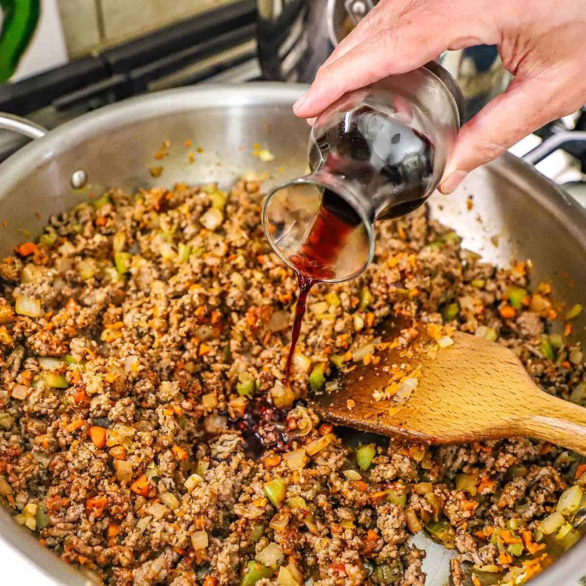 A person pouring red wine from a small glass carafe into a skillet filled with simmering ground beef and sausage with sautéed vegetables.