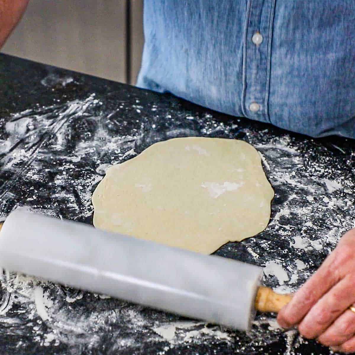 A person using a marble rolling pin to roll out a thin oval-shaped dough for homemade naan on a floured marble surface.