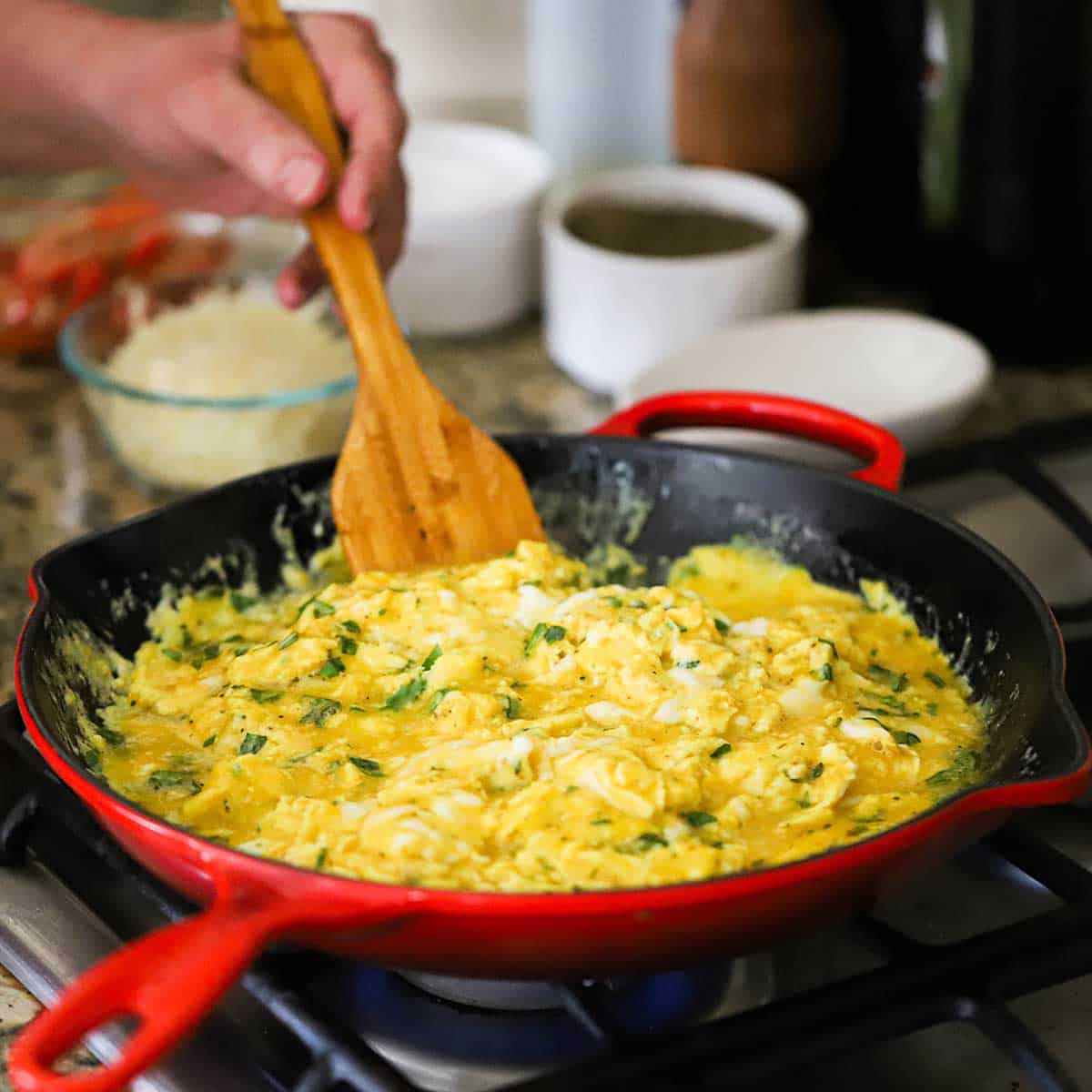 A person using a wooden spatula to stir fresh eggs that are being scrambled in a large skillet along with fresh basil on a gas stove.