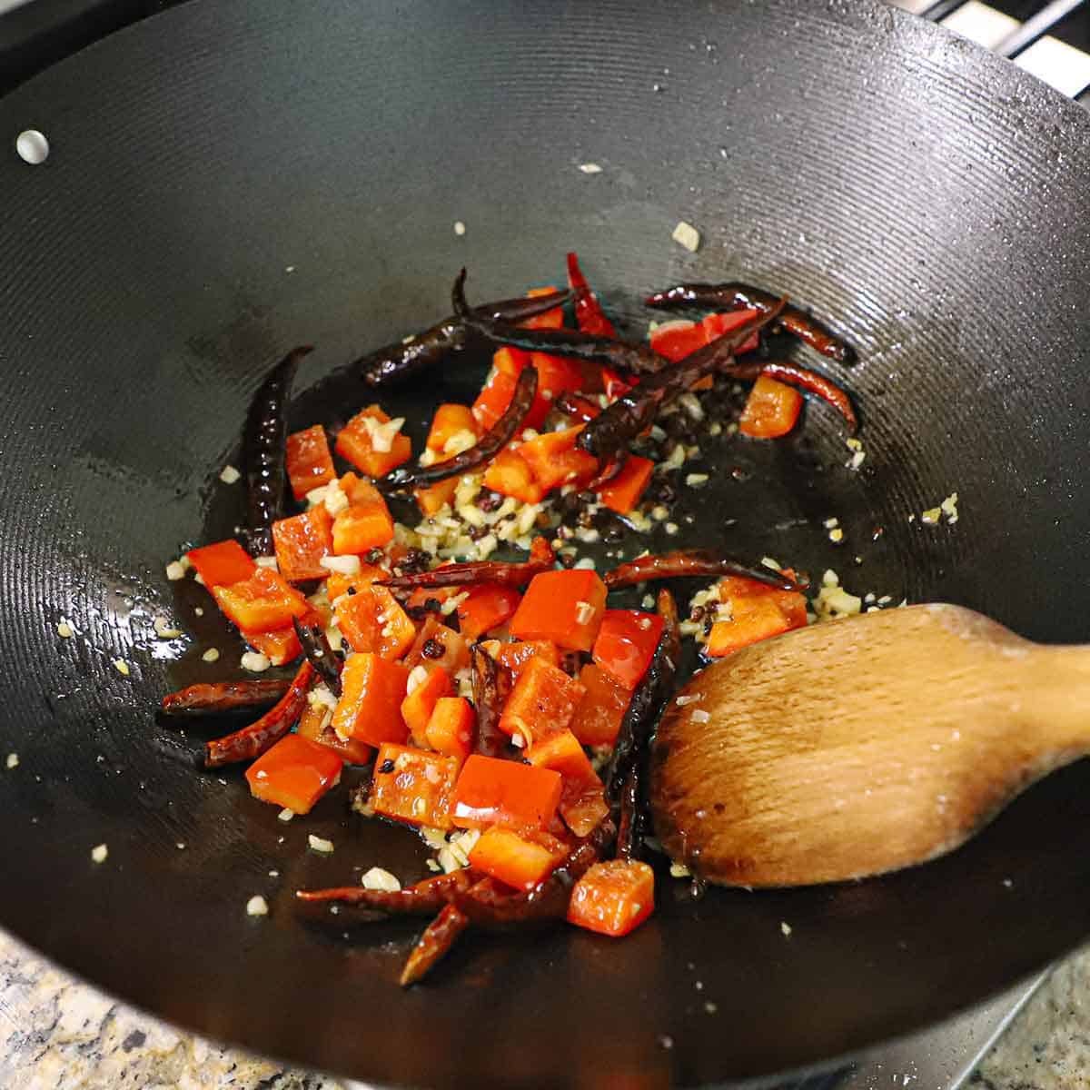 A person using a wooden spoon to stir-fry minced garlic and ginger, as well as chunks of red bell pepper and dried red chiles in hot oil in a large wok on a stovetop.