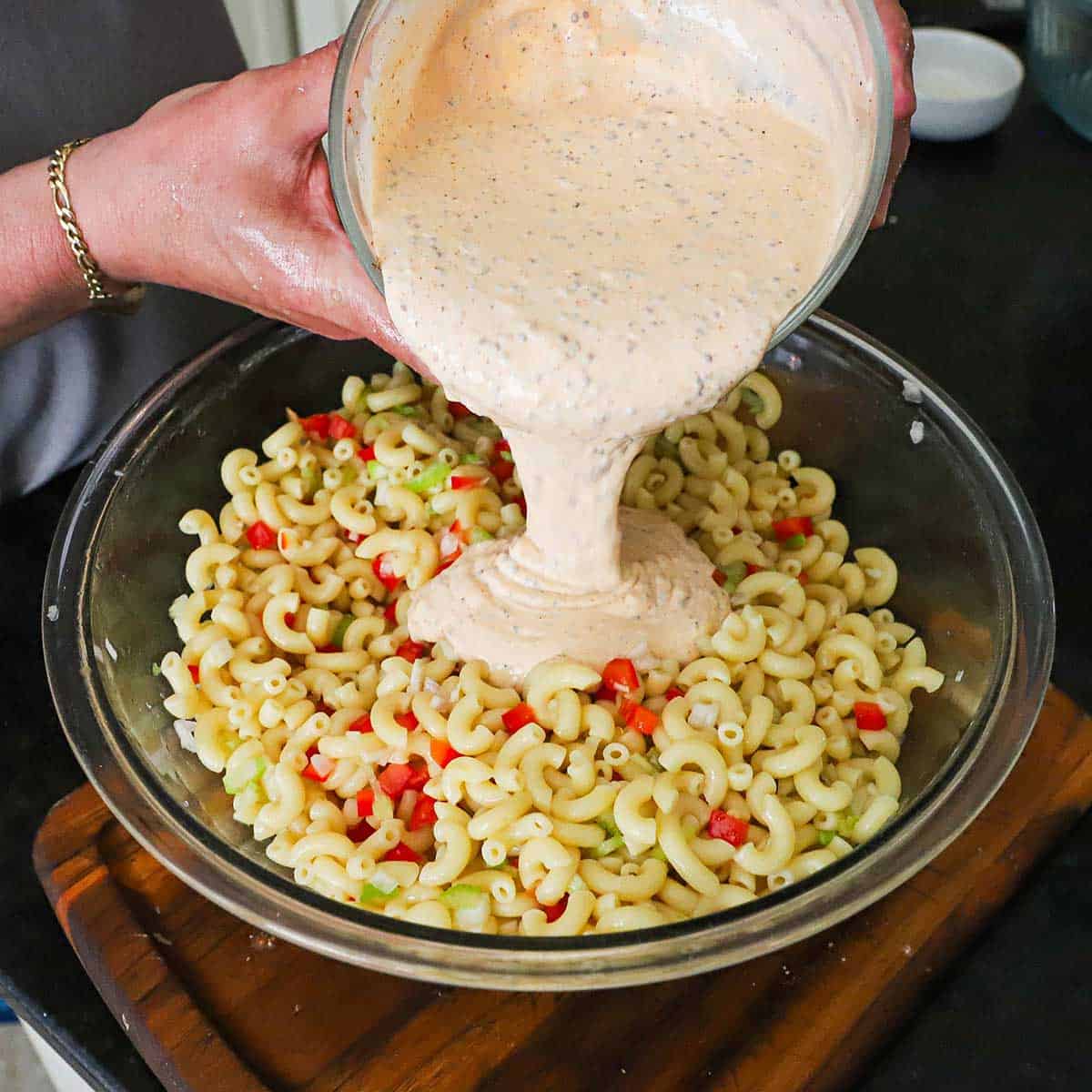 A person pouring a Cajun mayonnaise-based dressing into a large glass bowl that is filled with cooked macaroni pasta and chopped vegetables.